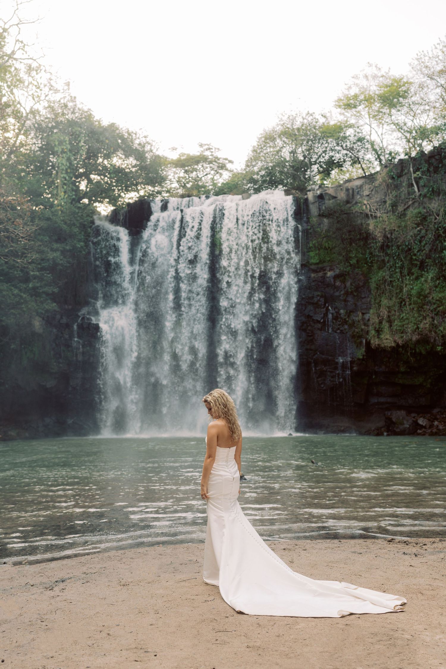 Wedding dress trailing on sandy beach with dramatic waterfall backdrop.