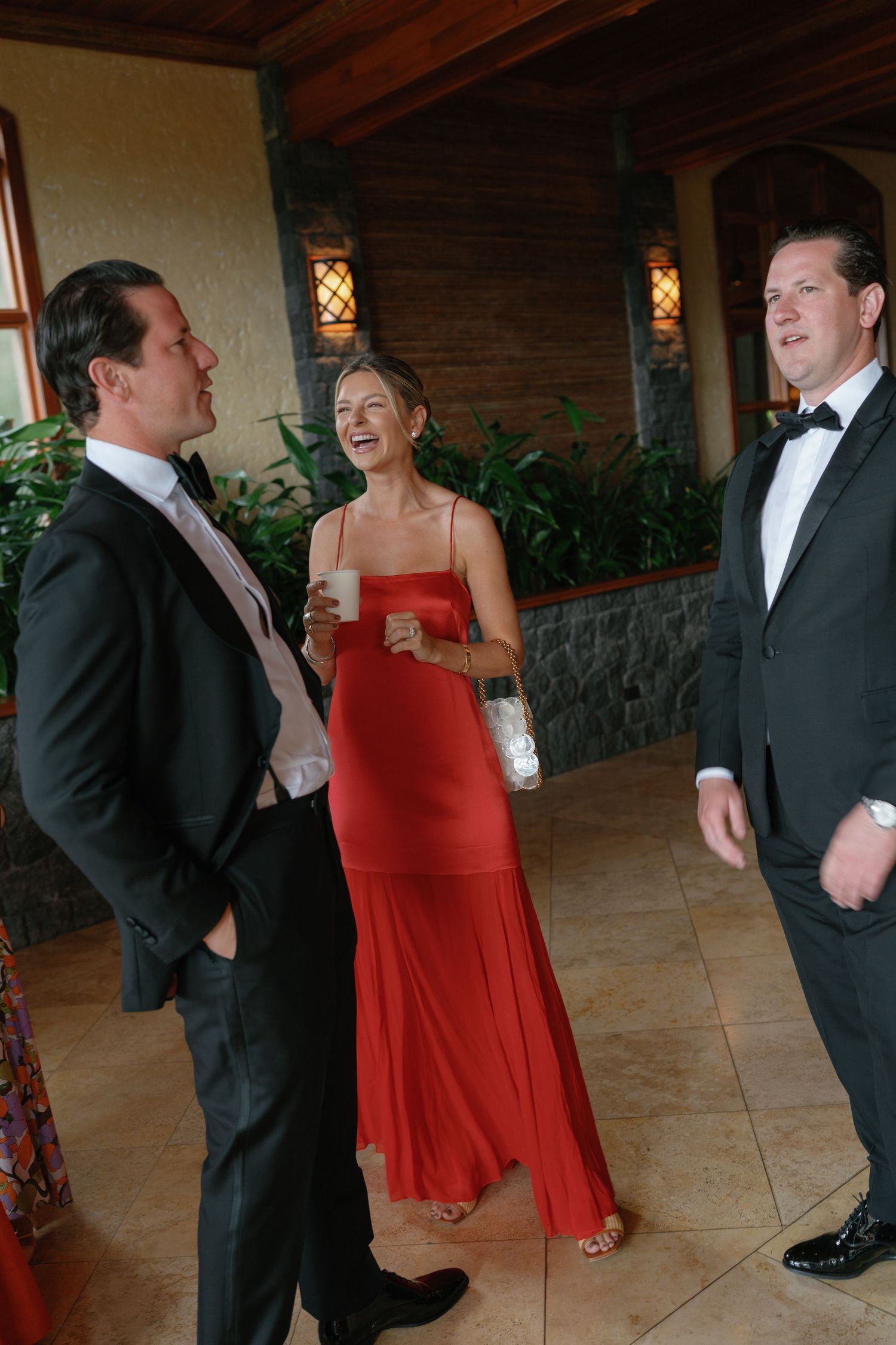 Formal event guests in black tuxedos and red dresses mingling under covered patio.