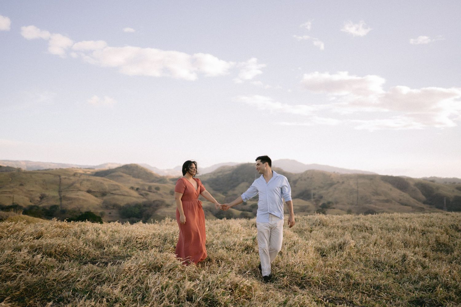 Couple walks hand in hand through rolling hills during golden hour.