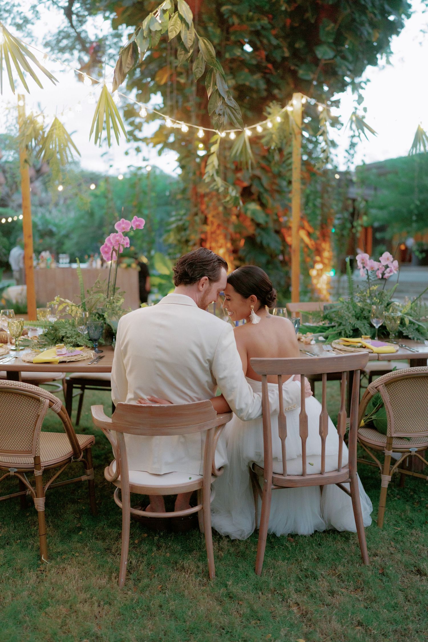 Romantic outdoor wedding reception setup with string lights and vintage chairs in a garden setting at dusk.