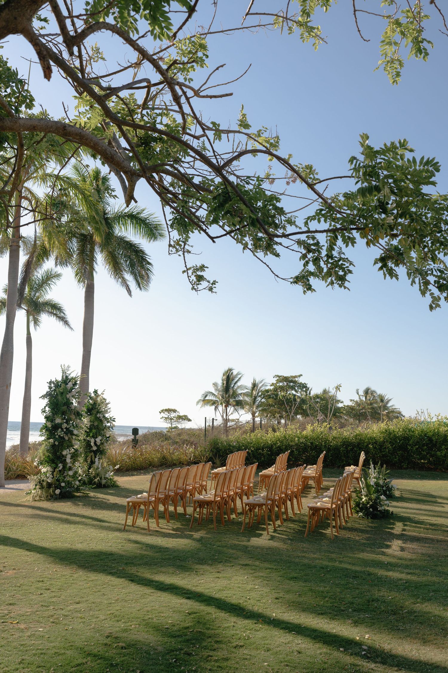 Outdoor wedding ceremony setup with wooden chairs on green lawn, flanked by floral arrangements and palm trees near the ocean.