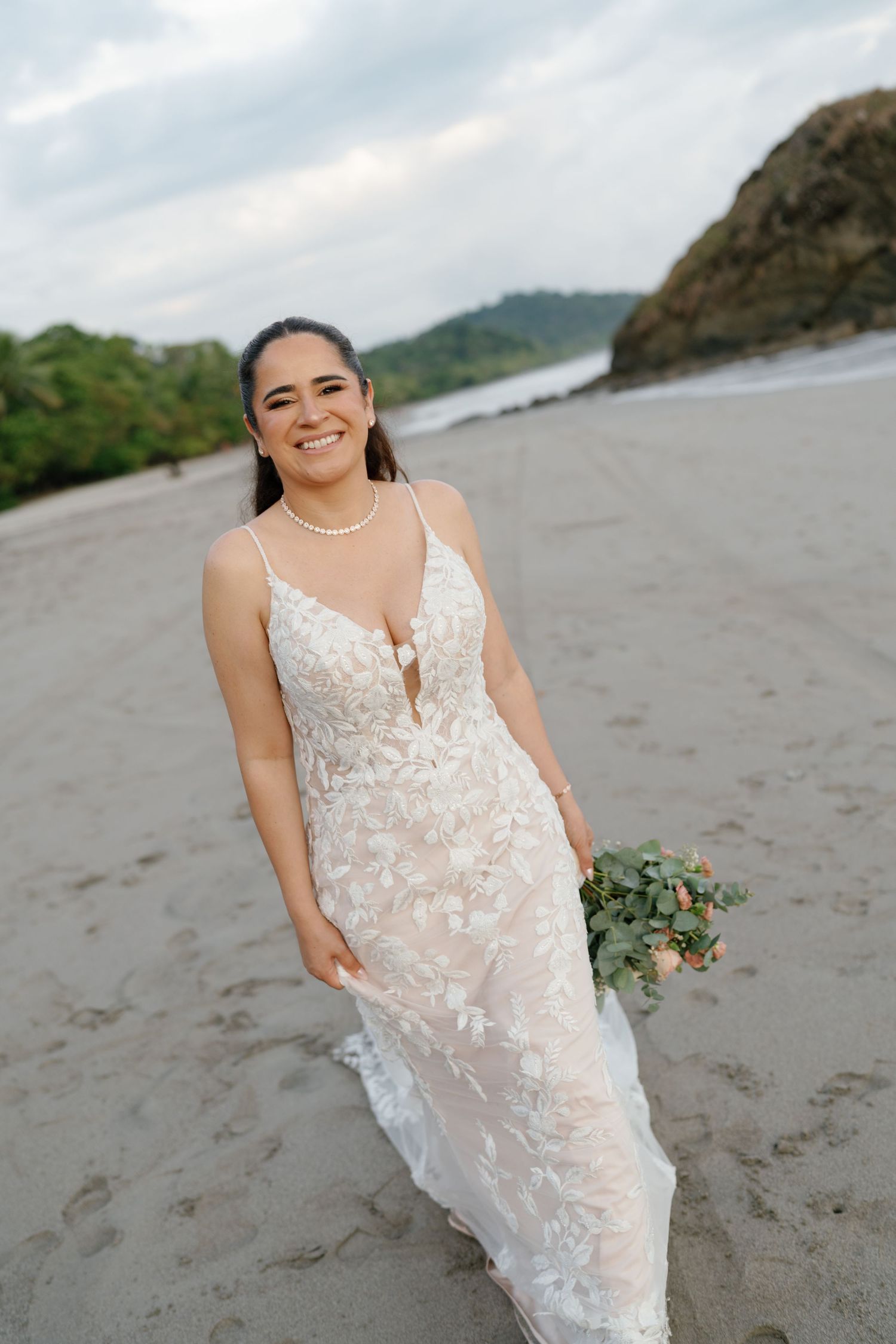Bride in lace wedding dress poses on beach with bouquet.