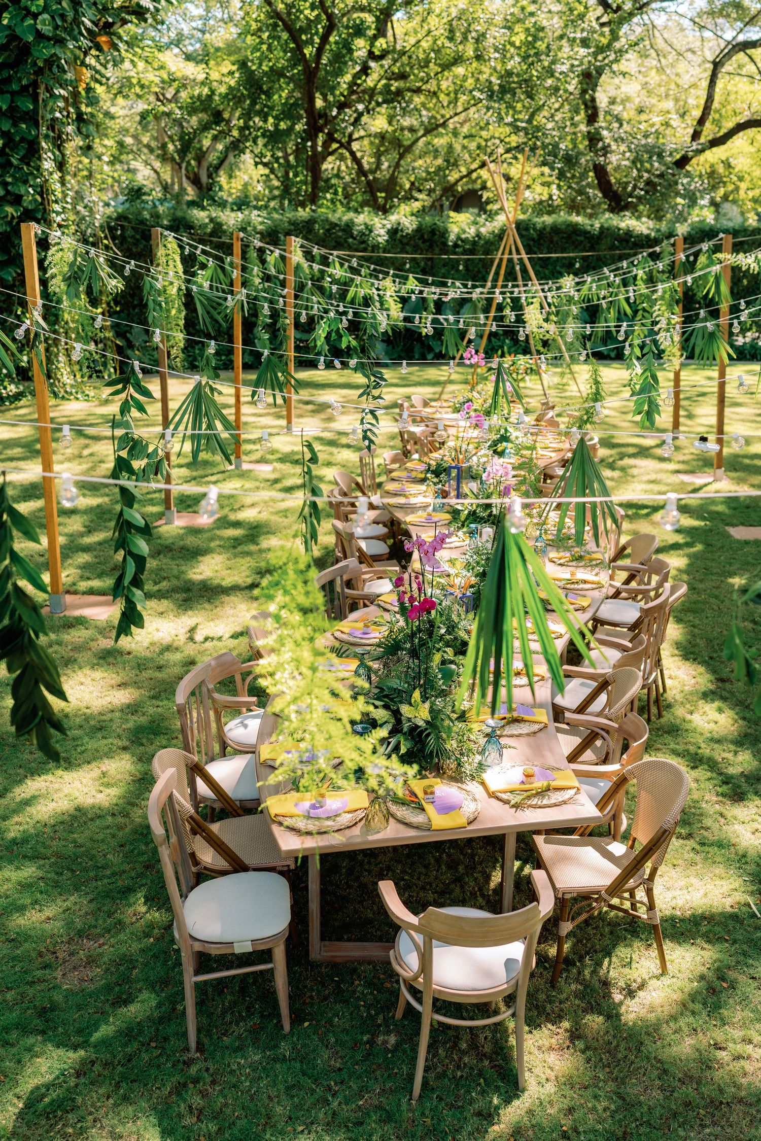 Garden wedding reception tables decorated with tropical greenery and string lights overhead.
