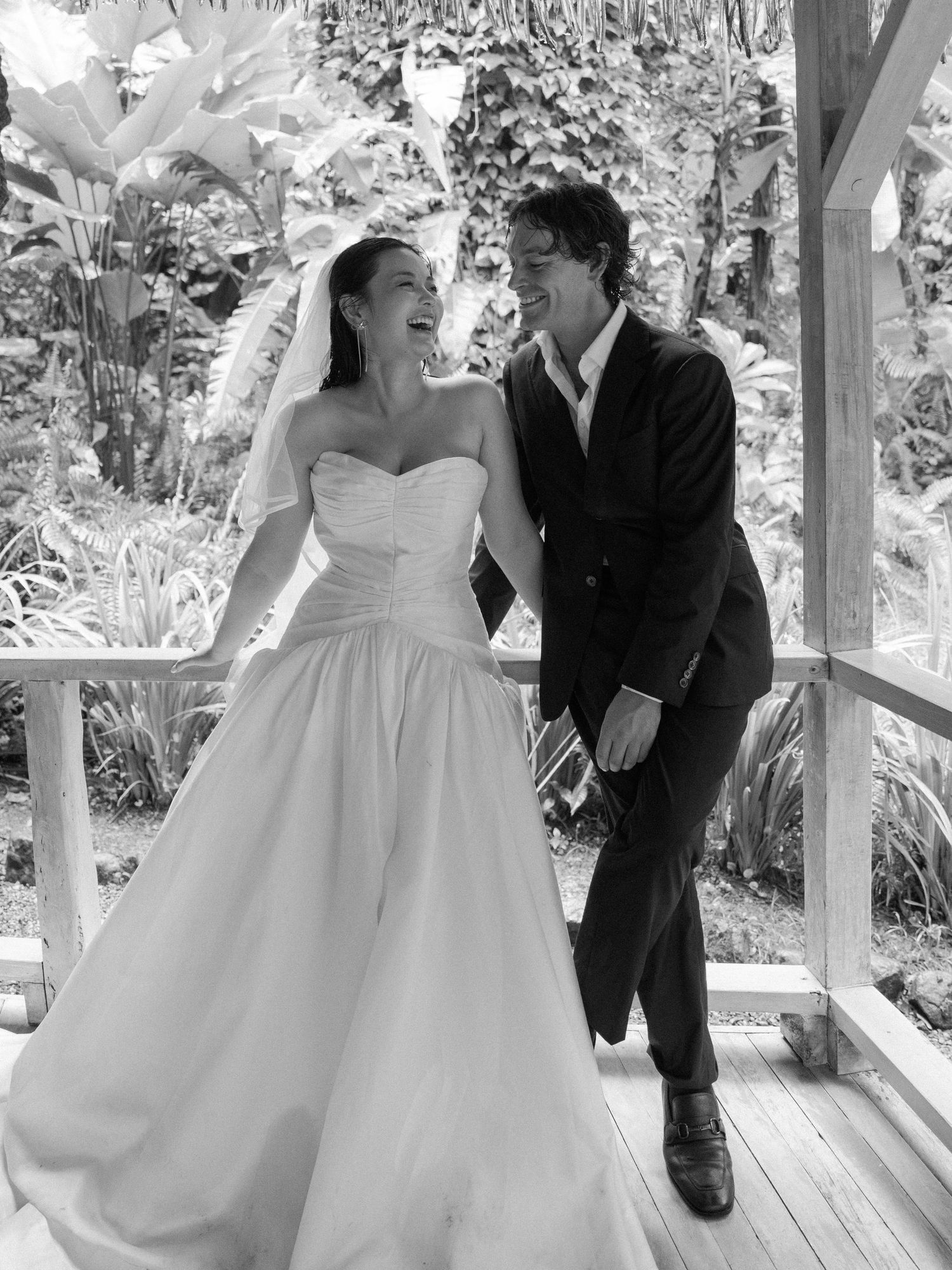 Joyful wedding couple sharing intimate moment on wooden porch surrounded by tropical plants.