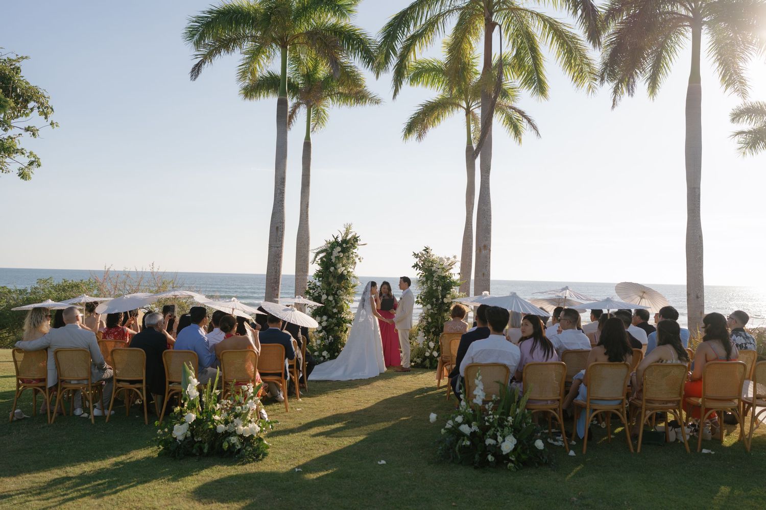 Outdoor beach wedding ceremony with palm trees and ocean views as guests sit in wooden chairs on a grassy lawn.