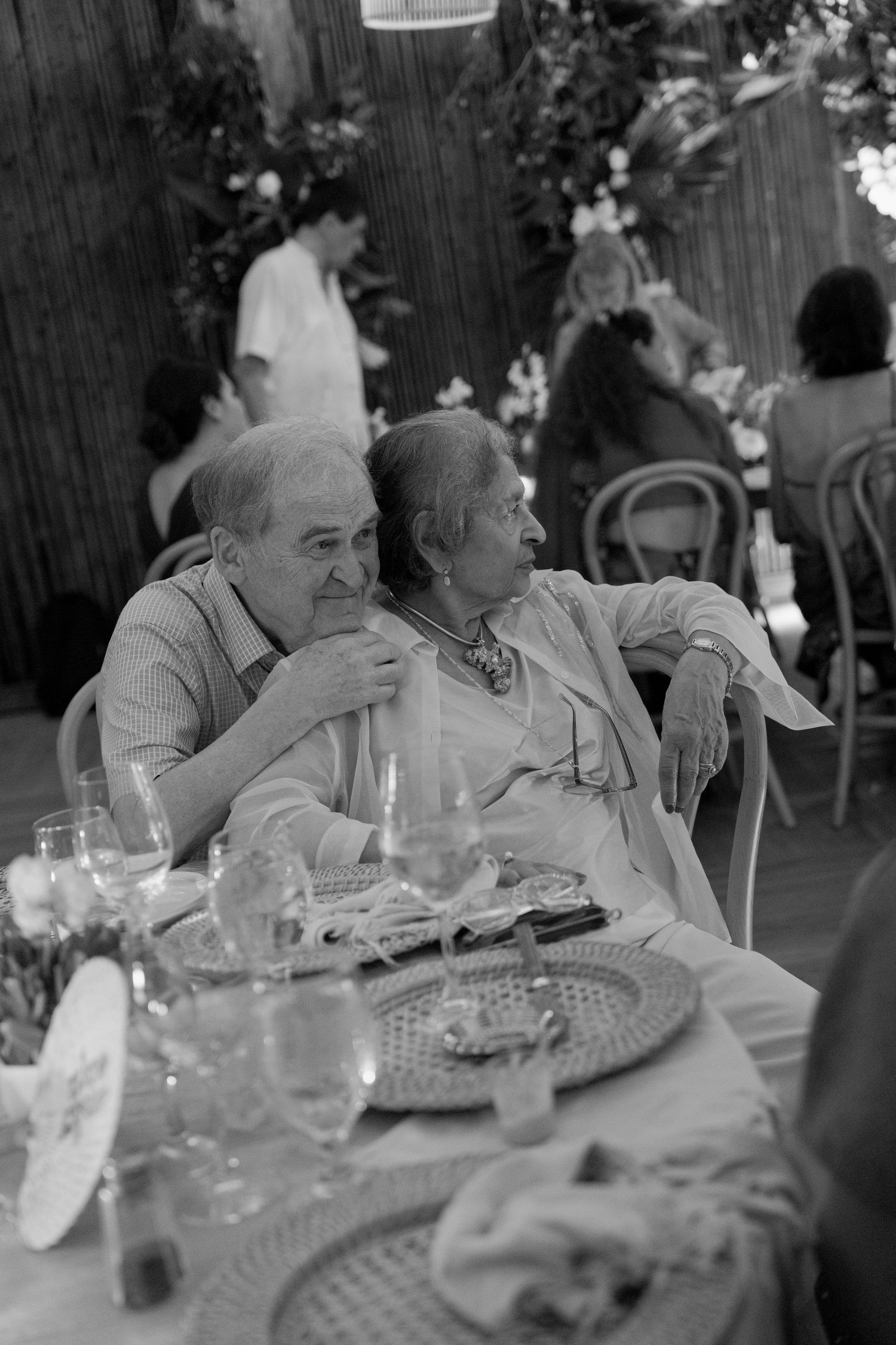 Two elderly people sit at an outdoor dining table with plates and glasses, engaged in conversation at a social gathering.