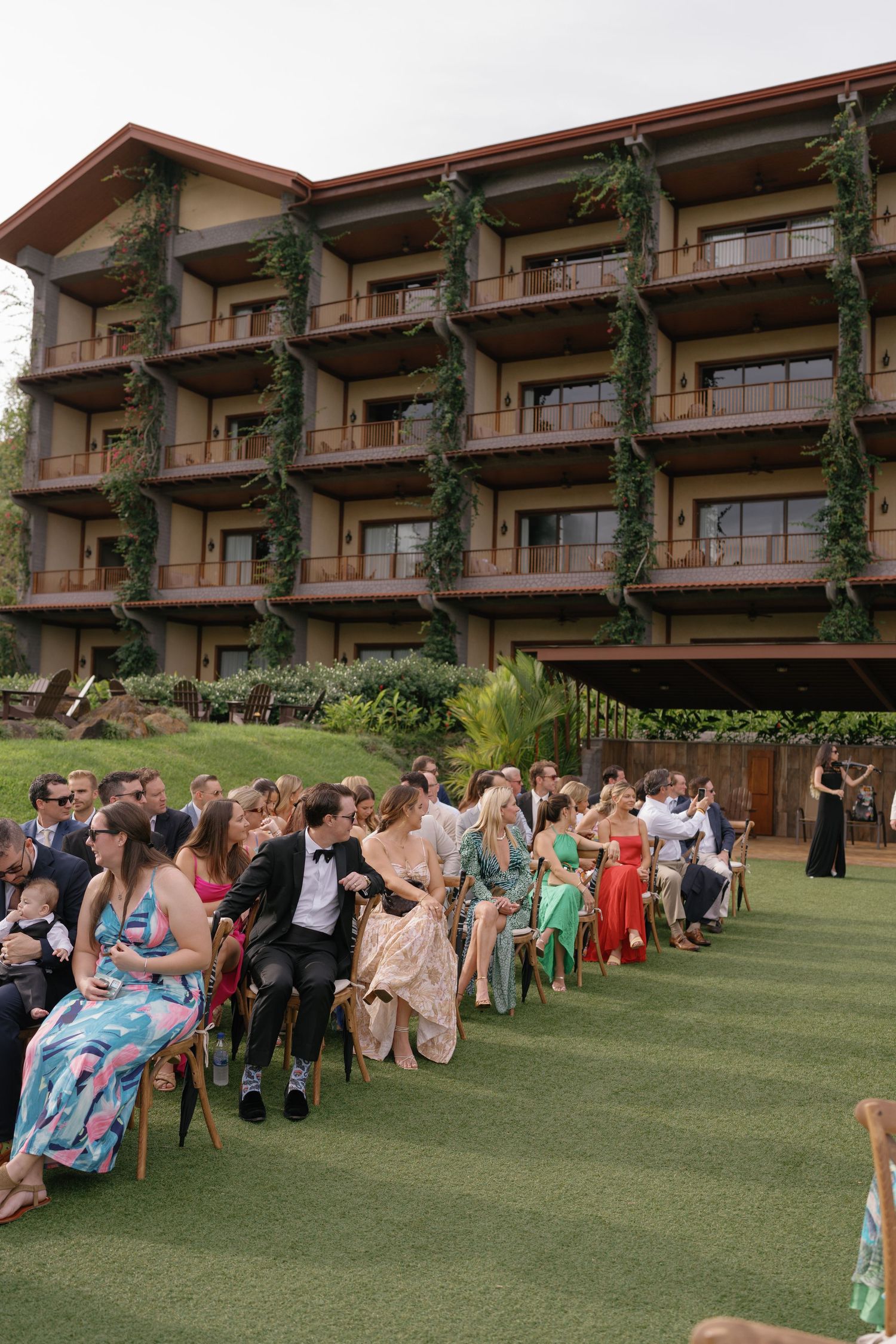 Wedding ceremony guests seated on lawn in front of modern resort building.