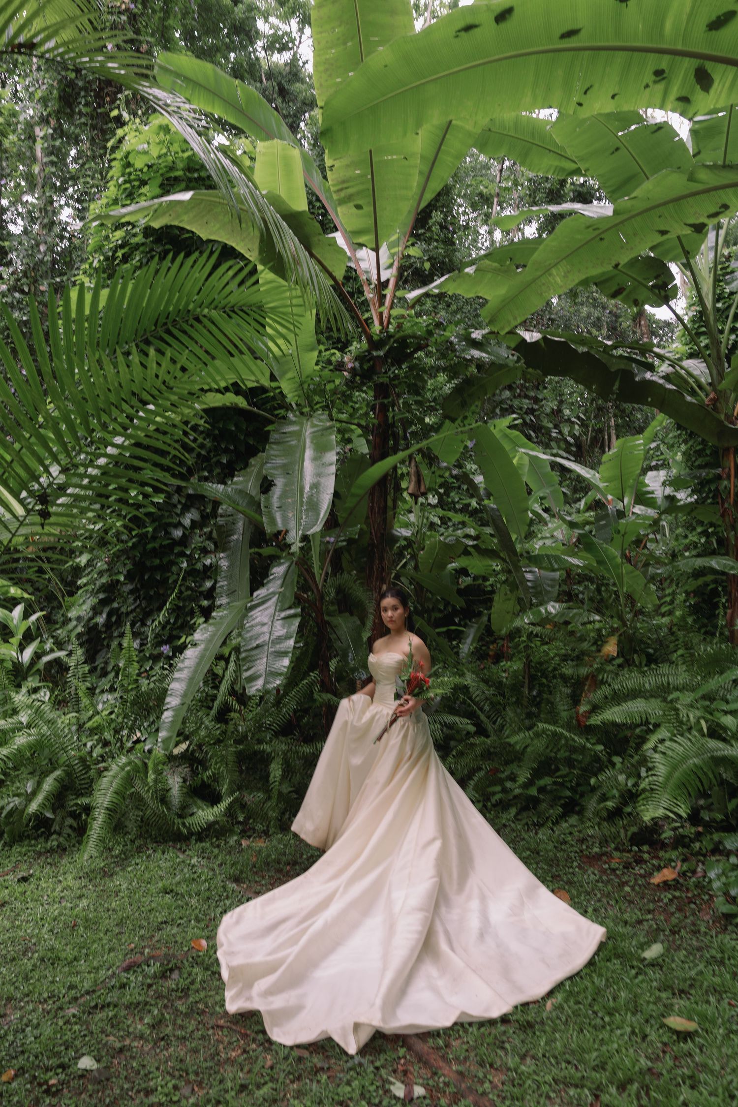 A bride in a flowing white gown poses elegantly among tropical banana leaves and ferns.
