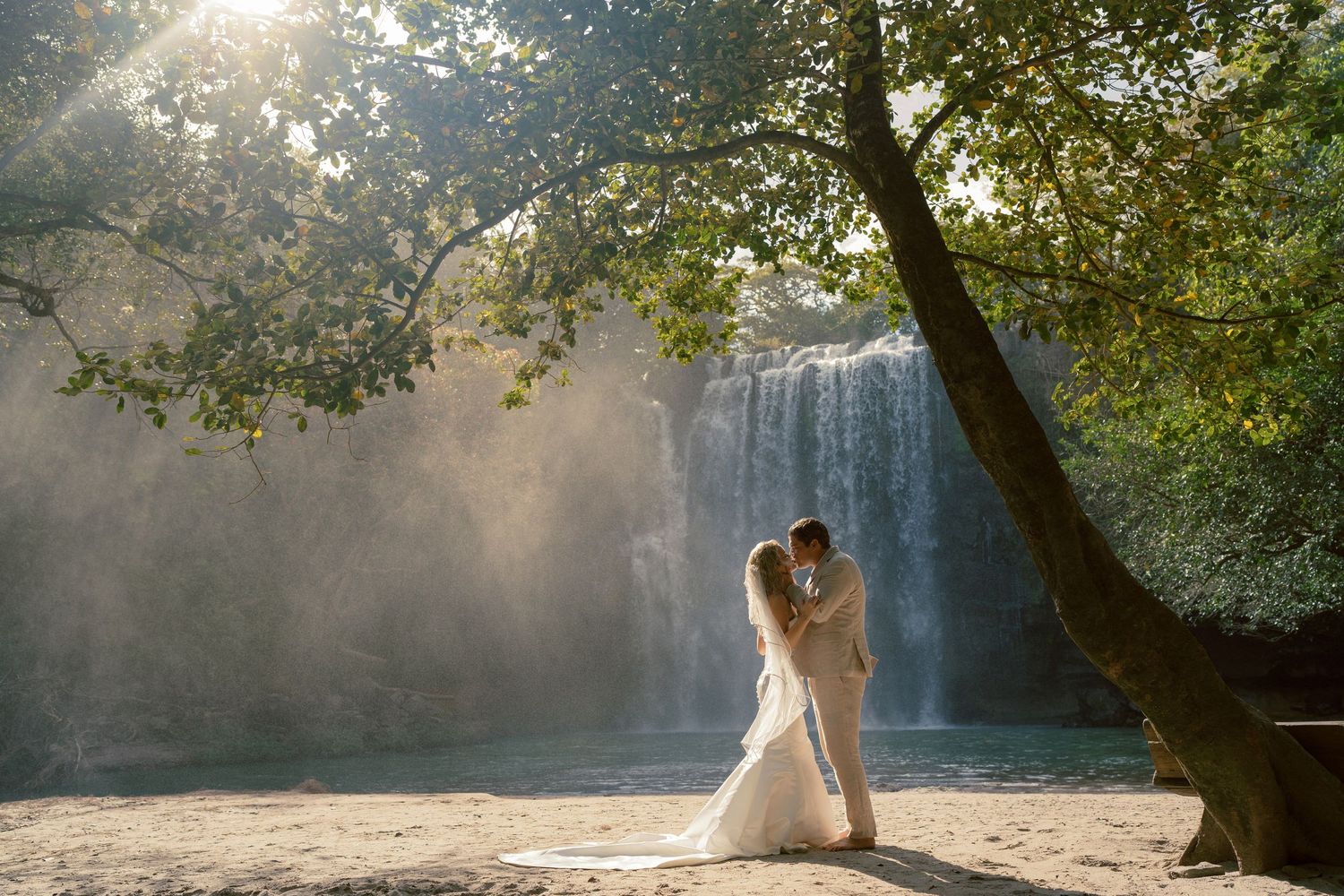 Couple posing by a scenic waterfall with sunlight streaming through trees.