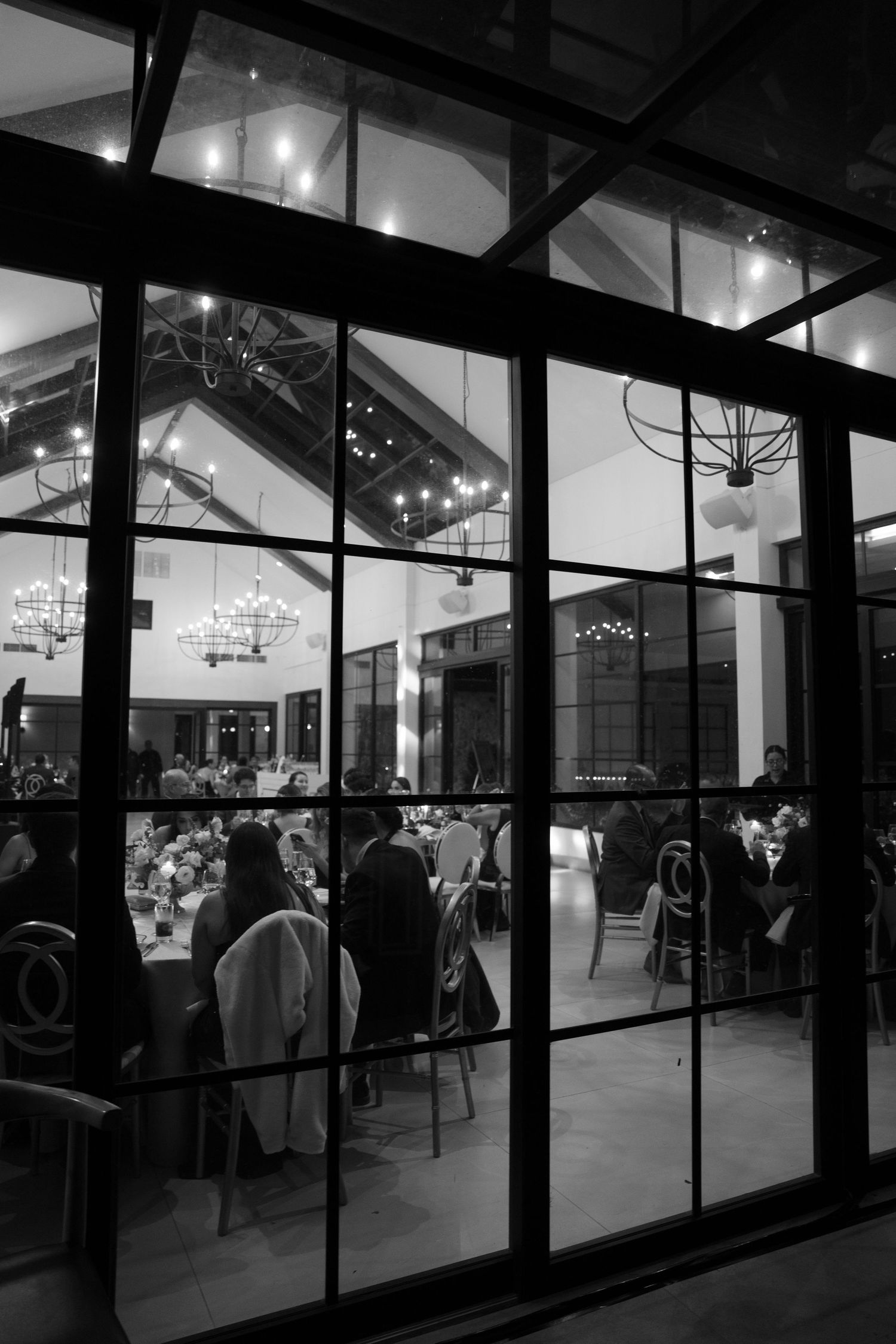Black and white panoramic view of people silhouetted through large glass windows in a modern building at night.