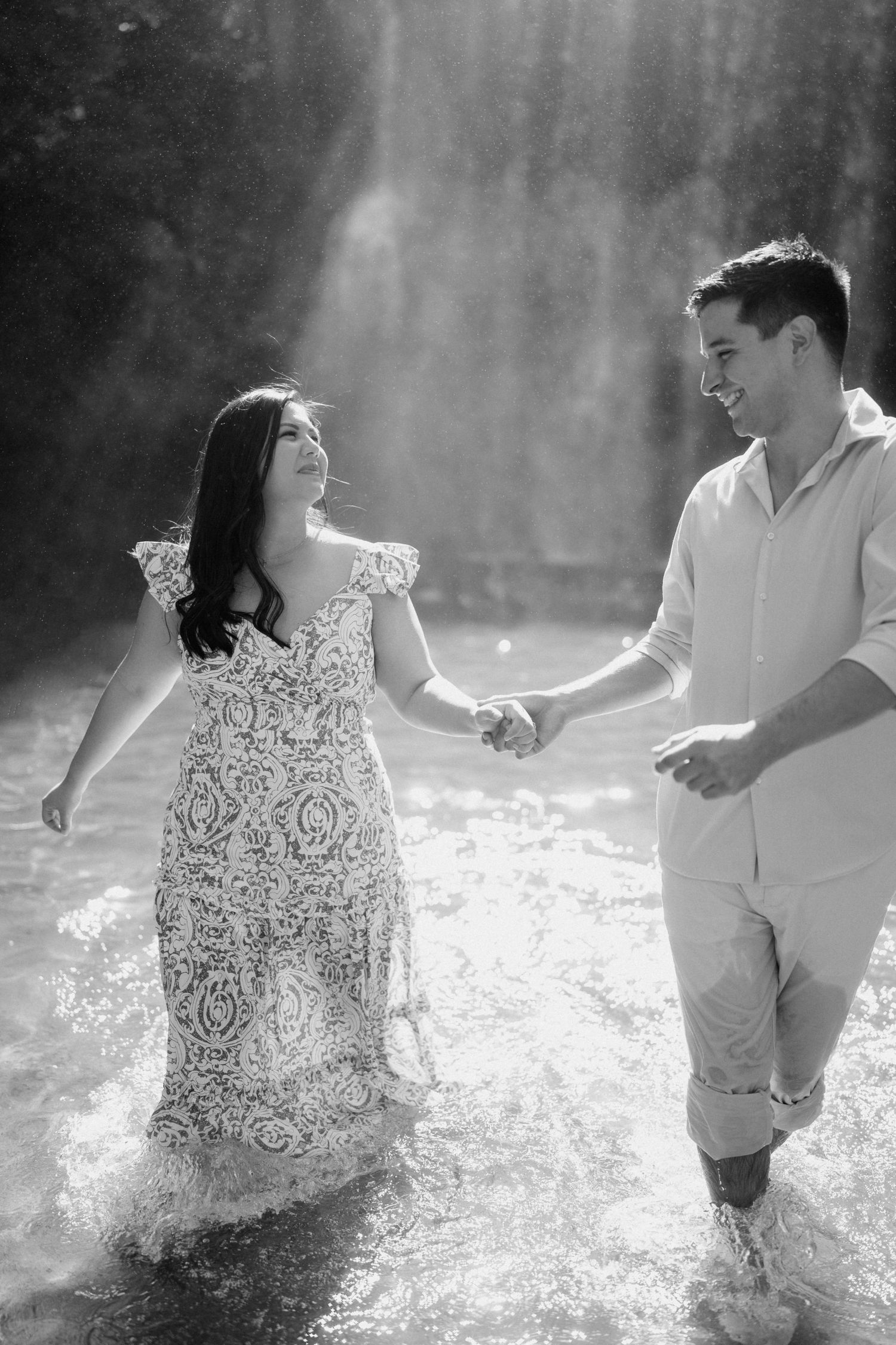 A couple splashes joyfully in shallow water during a black and white photoshoot.