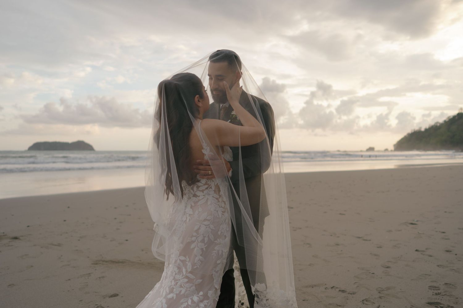 A couple embraces on a sandy beach with cloudy skies and ocean in the background.