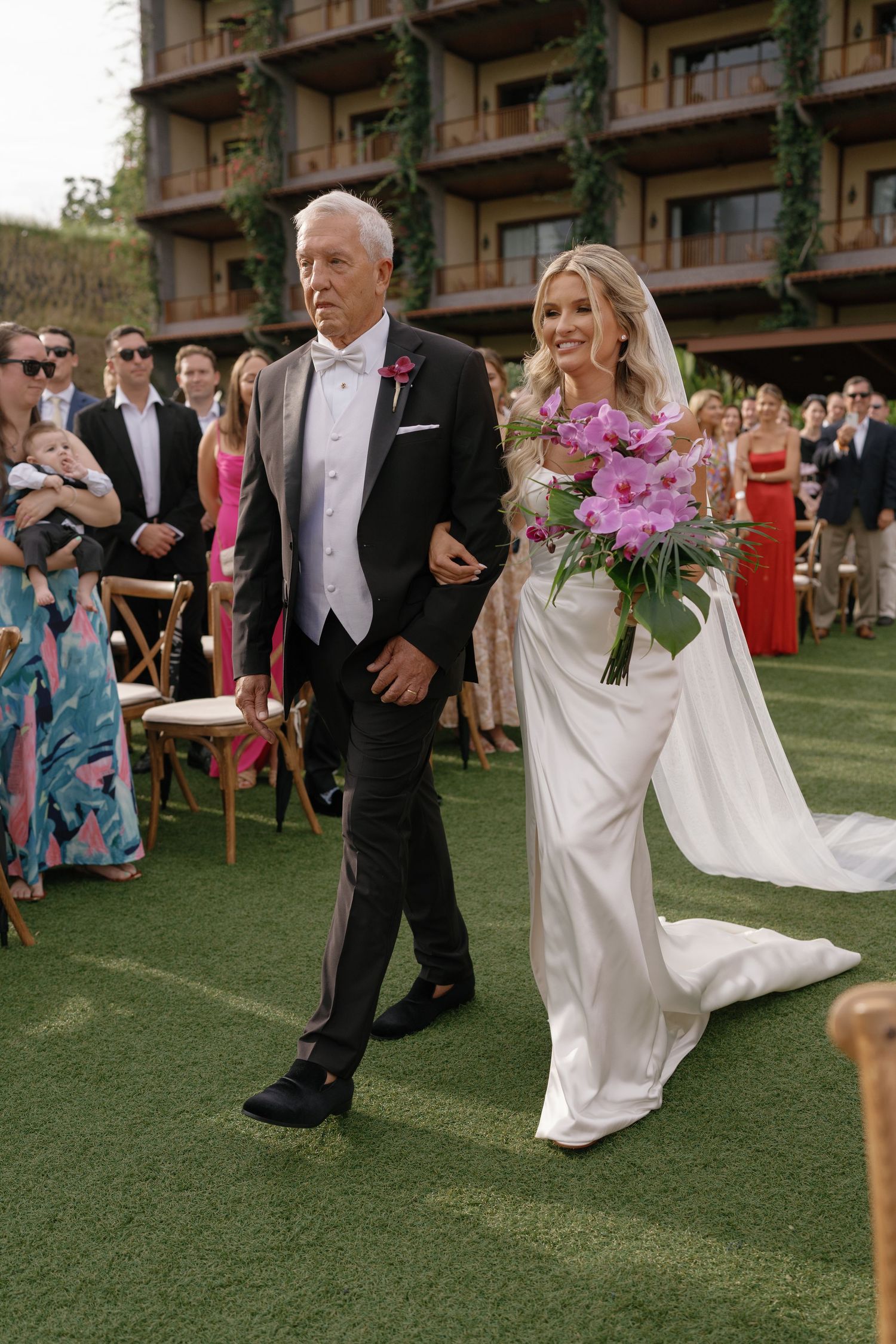 Wedding party walks across manicured lawn in front of resort building with pink flowers.