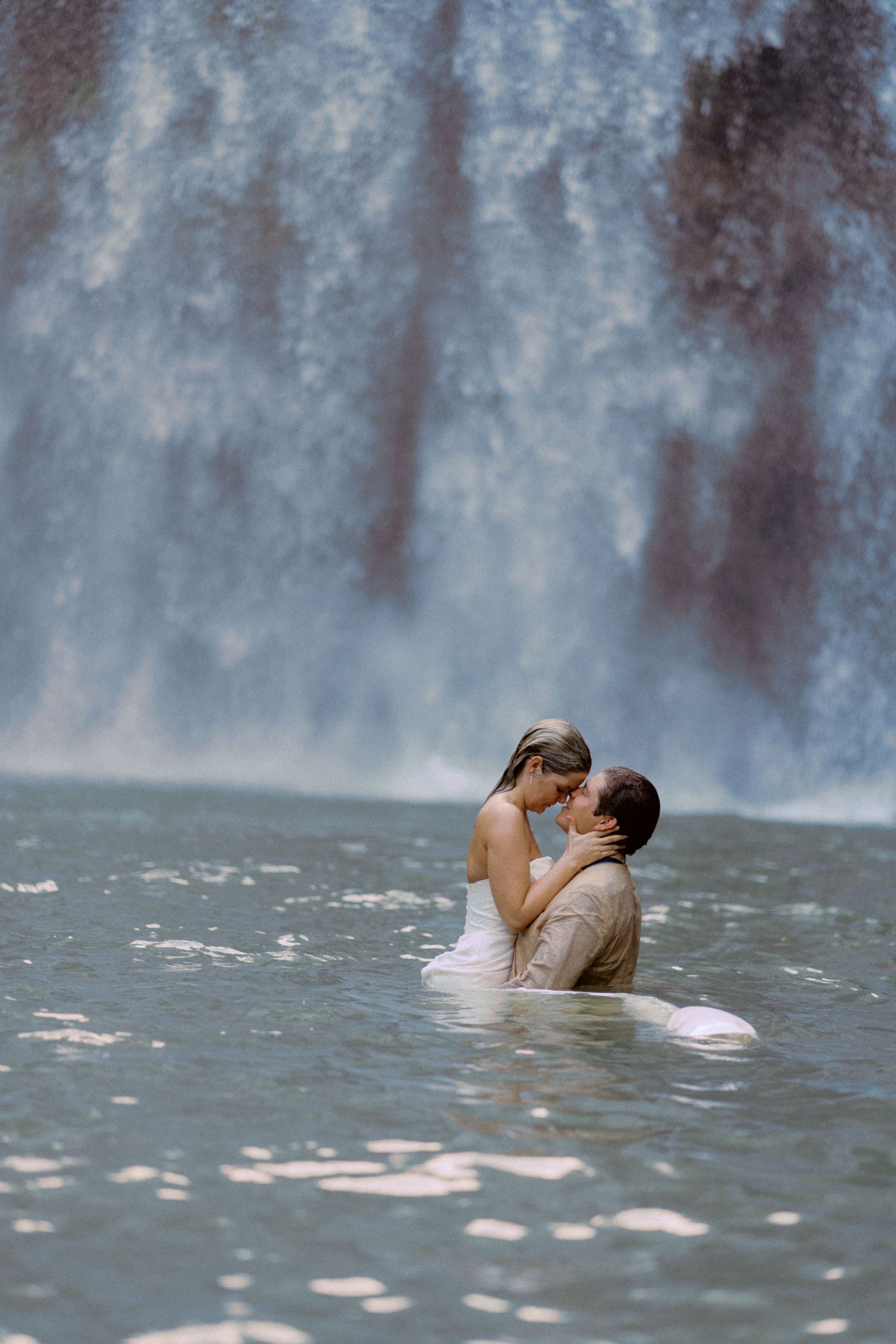 Two people embrace while swimming in the misty waters beneath a grand waterfall.