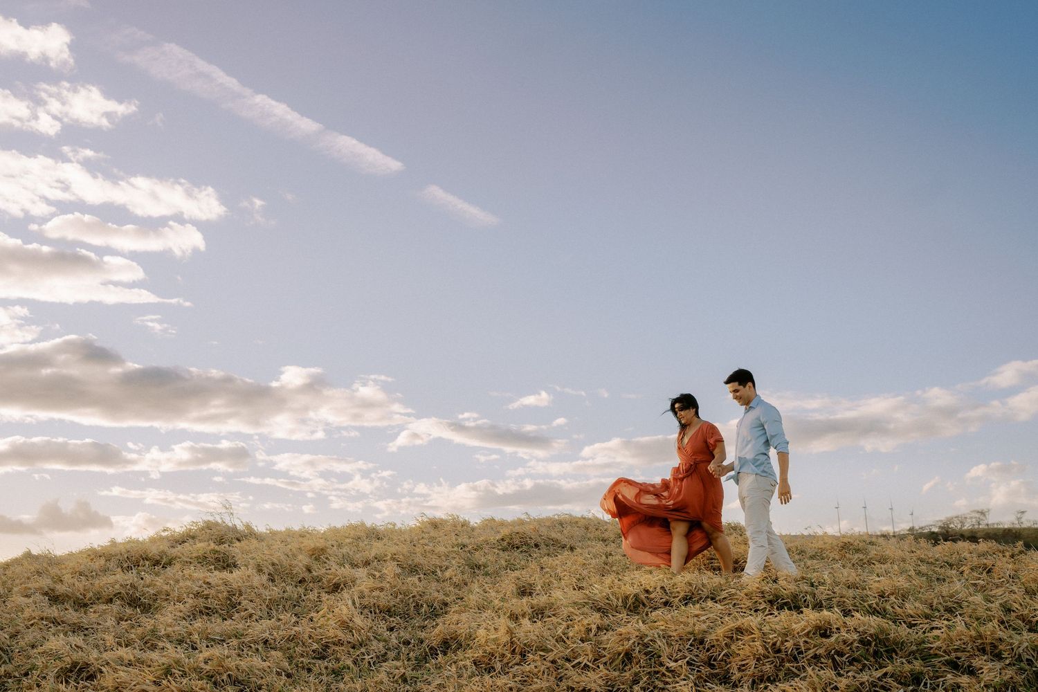 Two people stand together on a hilltop against a bright blue sky with wispy clouds.