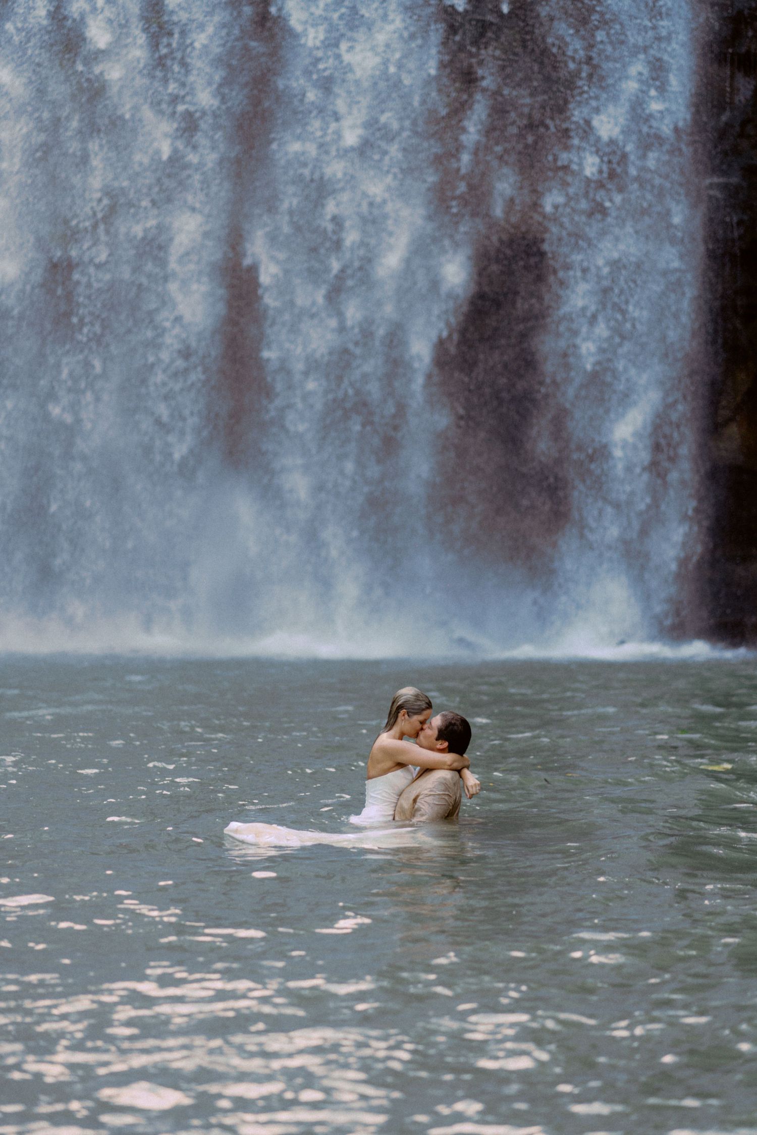 Two people share an intimate moment while swimming in front of a cascading waterfall.