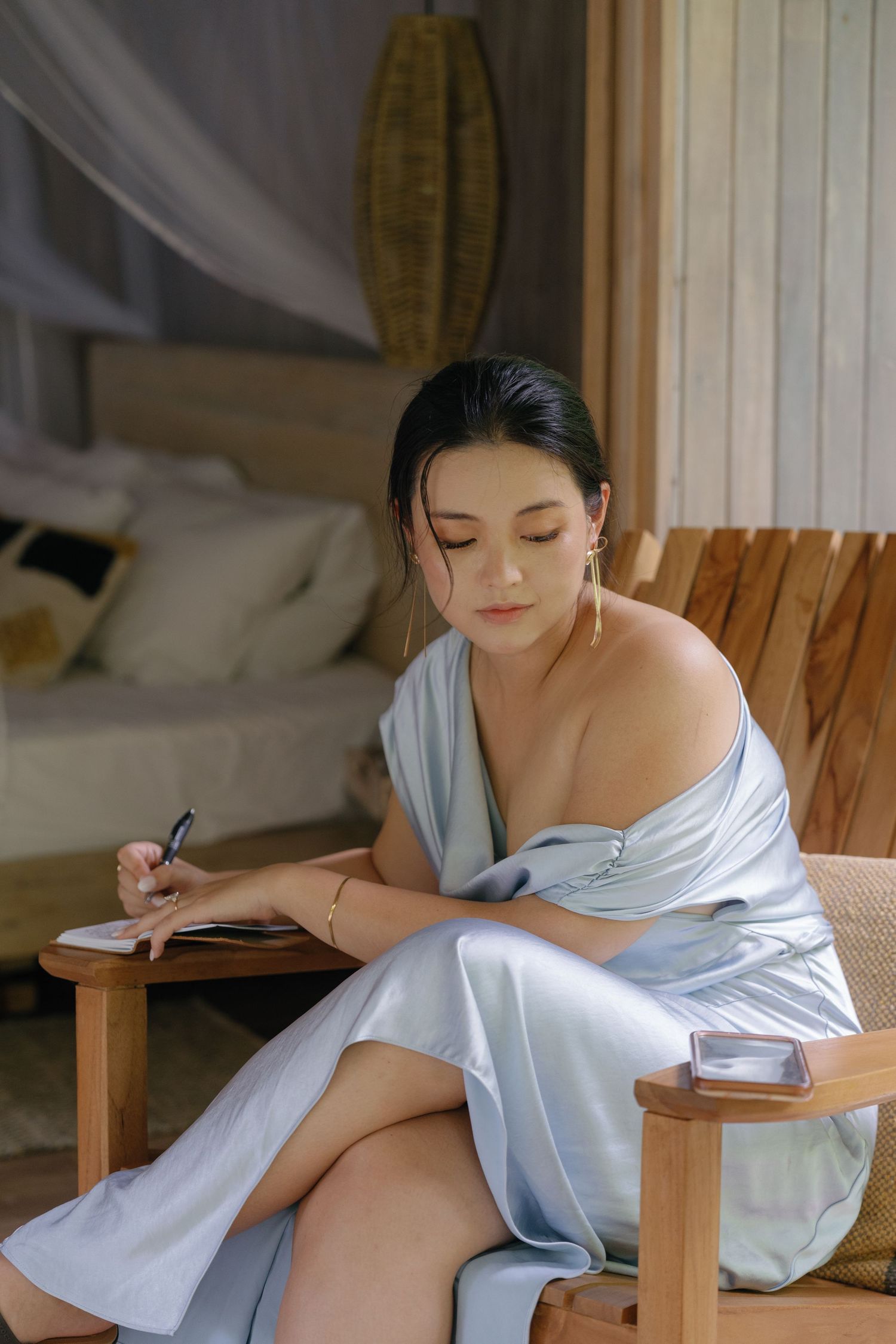Woman in light blue dress writing while seated in wooden chair with tropical decor in background.