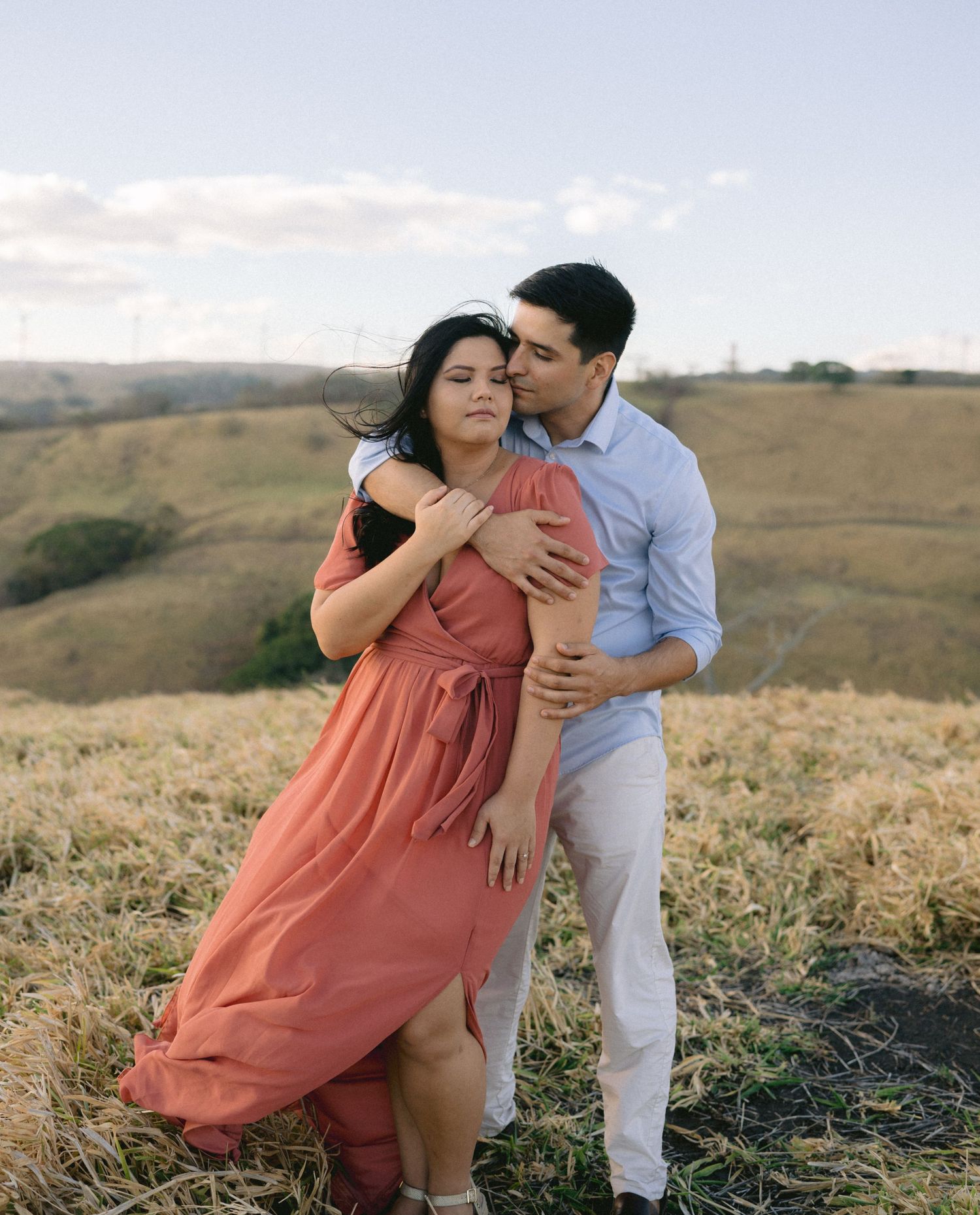 A couple shares a romantic dance pose in a coral dress and white outfit on a grassy hillside.