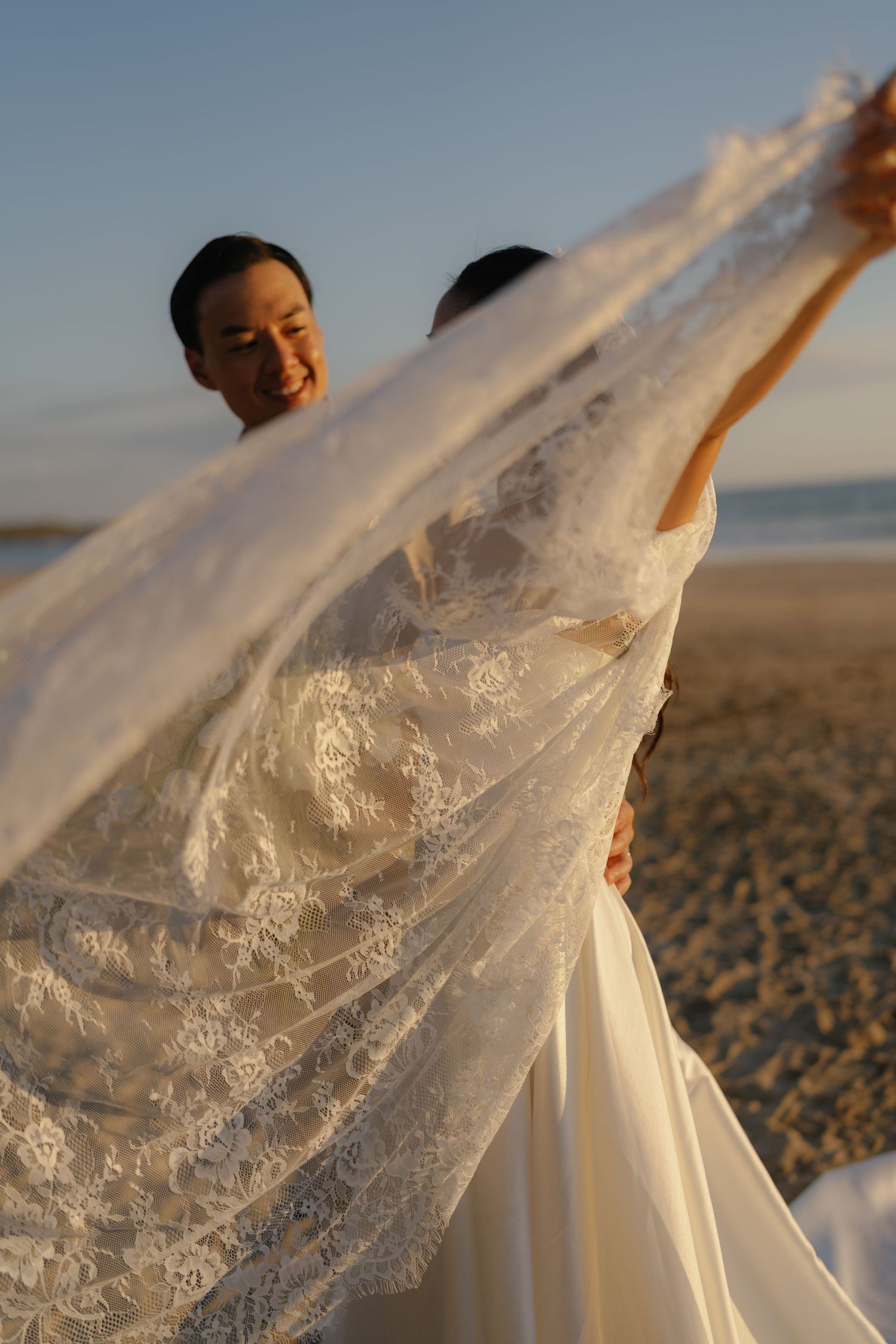 A white lace wedding dress flows elegantly in the wind against a beach backdrop during golden hour.