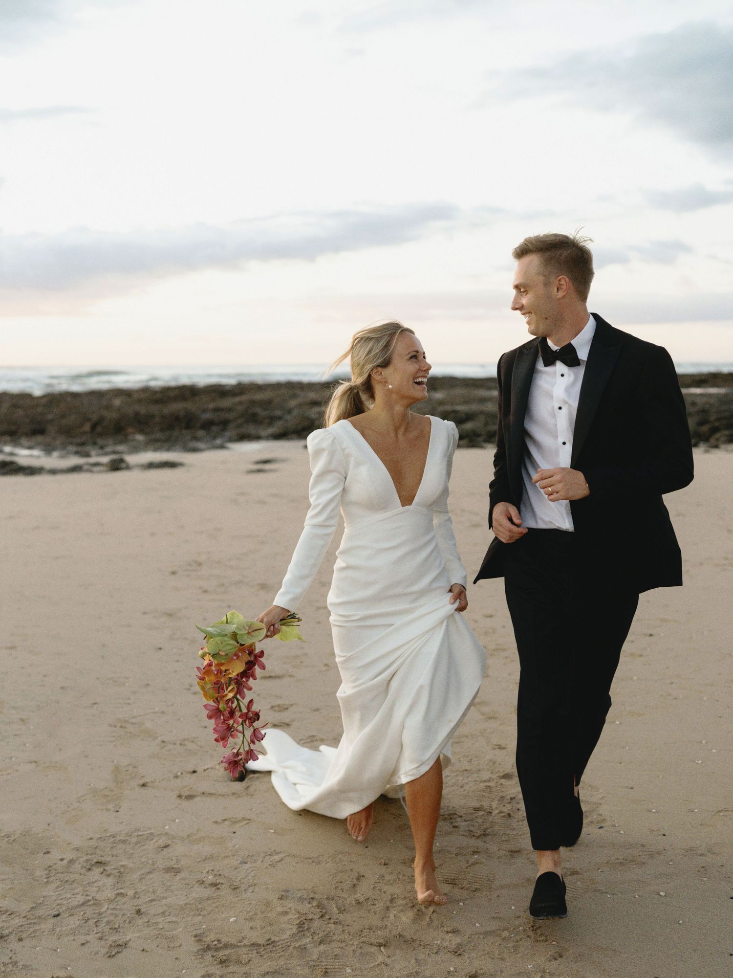 A couple in wedding attire runs playfully along a sandy beach at sunset, with the bride holding a vibrant bouquet.