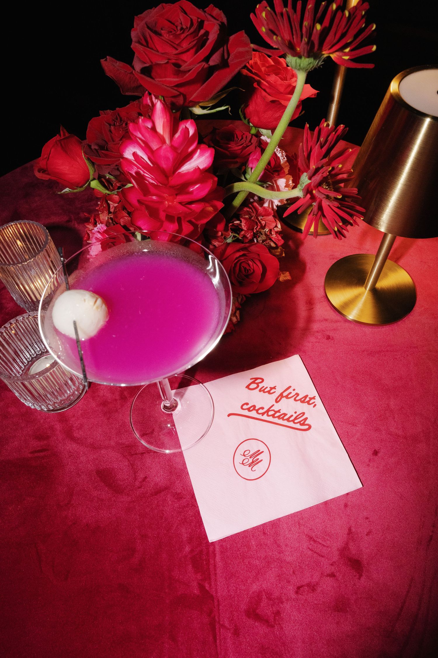 Pink cocktail with lychee garnish on a red velvet table with roses and a napkin reading 'But first, cocktails'.