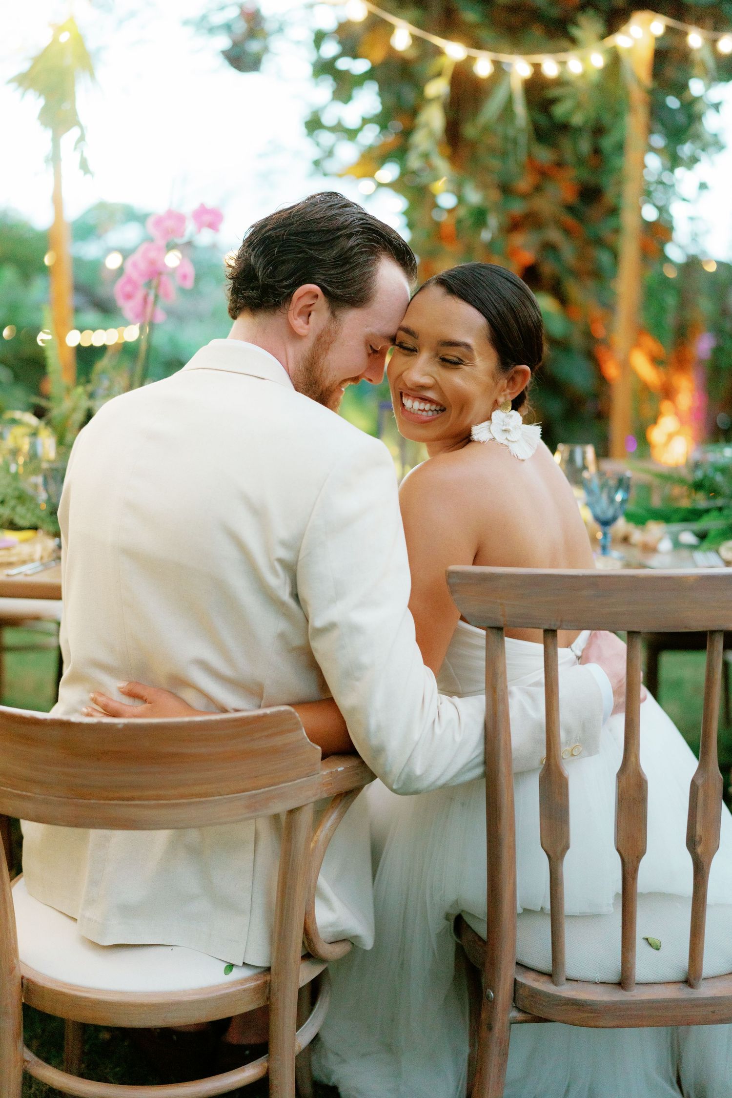 Wedding couple sharing tender moments at rustic outdoor reception with wooden chairs.