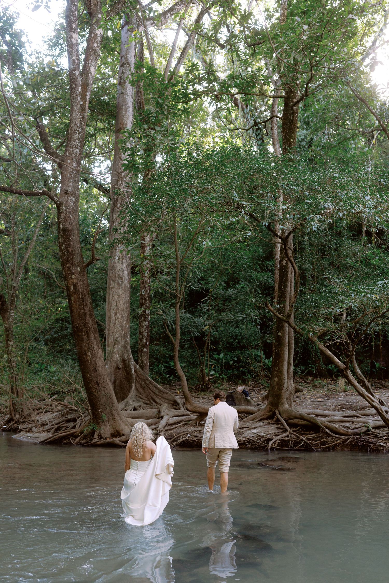 Couple wading into water wearing wedding attire near wooded riverbank.