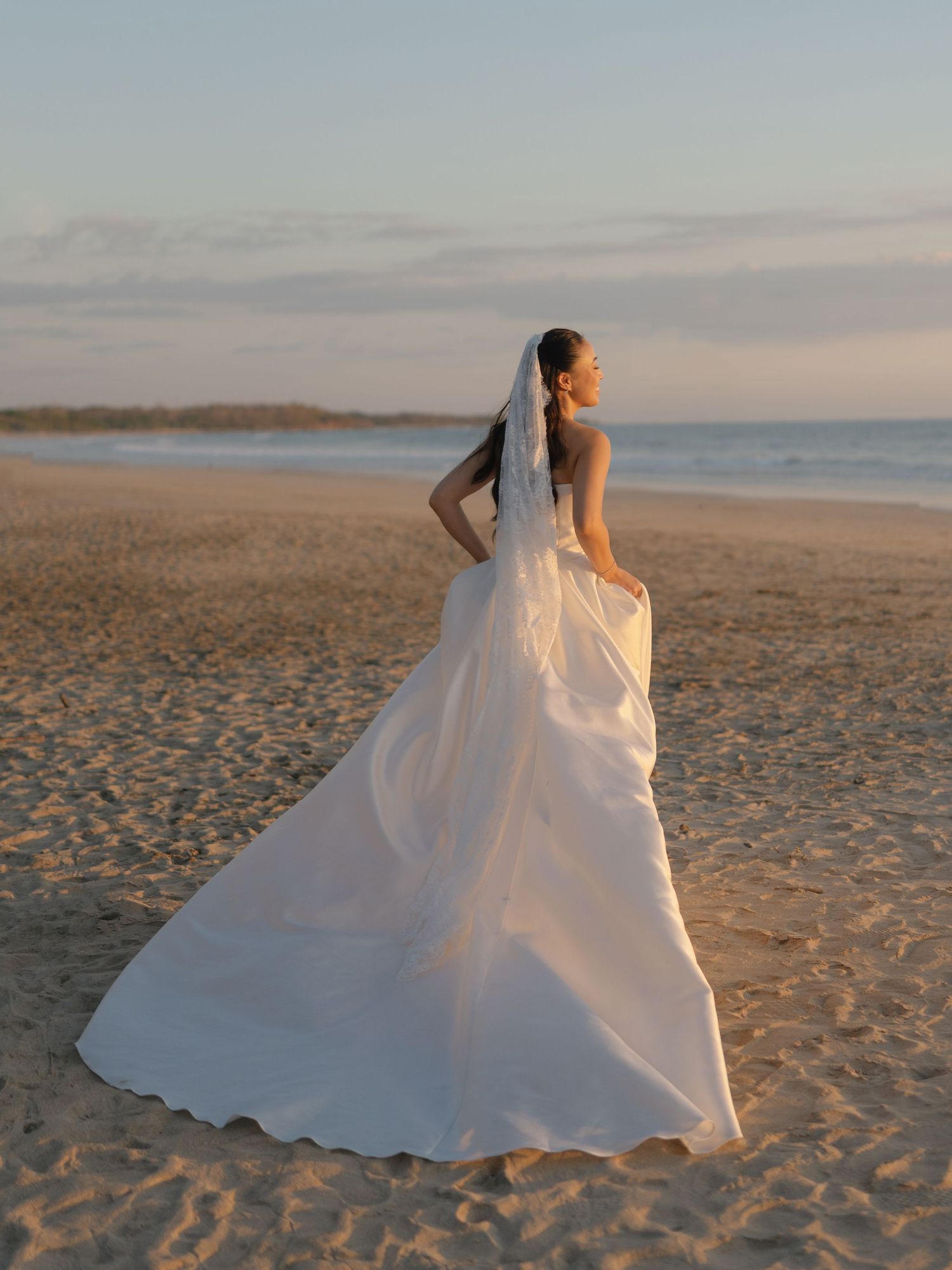 A bride in a flowing white ballgown twirls on a sandy beach during sunset.