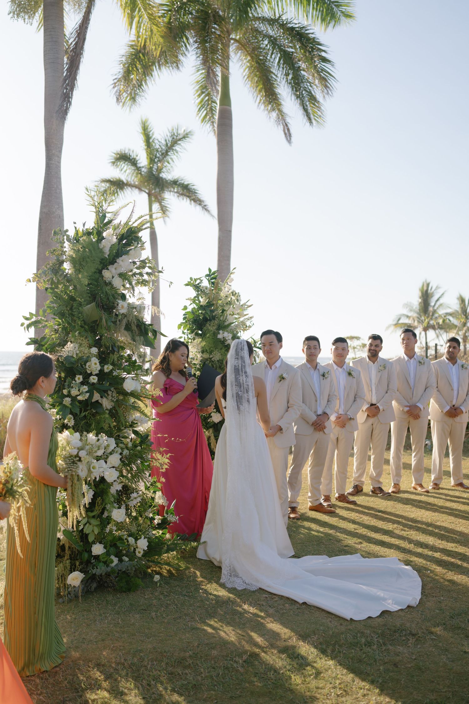 Outdoor wedding ceremony setup with palm trees and ocean backdrop featuring floral arrangements and white chairs.