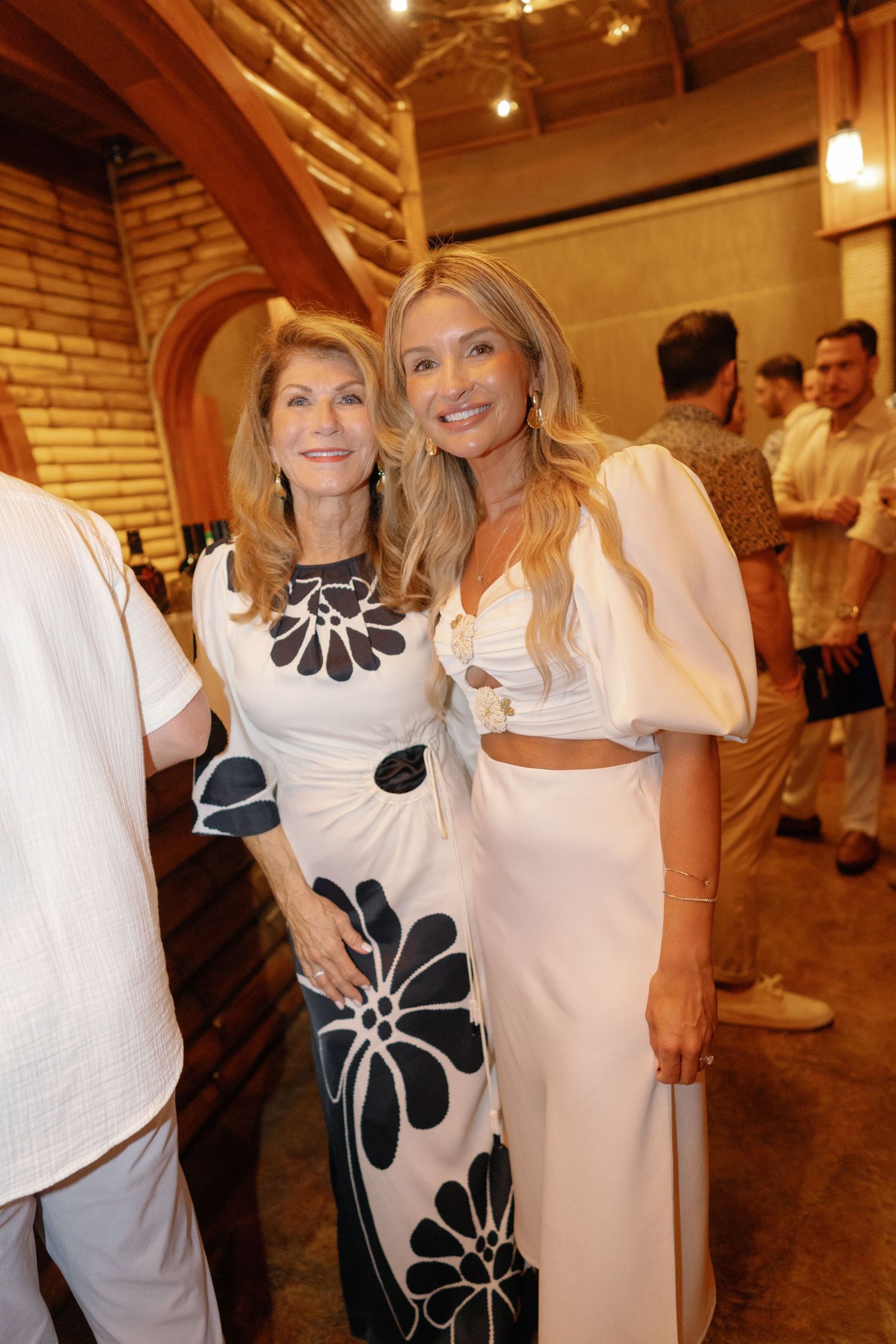 Two women in elegant white dresses with black floral accents pose together at an indoor event.