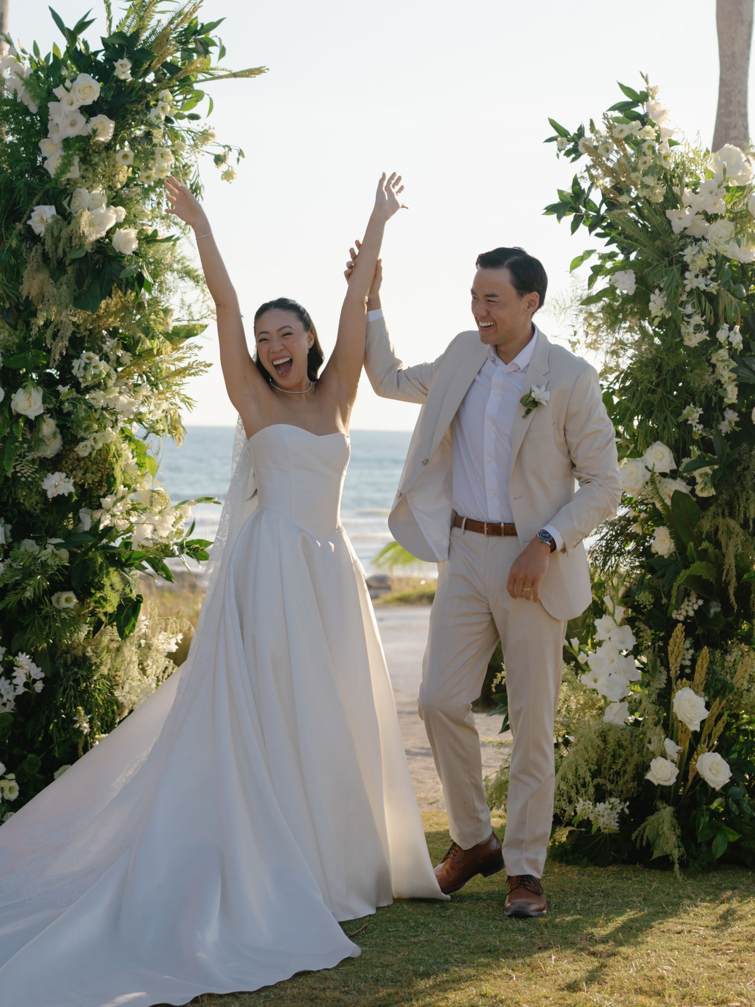A couple in white wedding attire dances and celebrates joyfully against a scenic outdoor backdrop with floral arrangements.