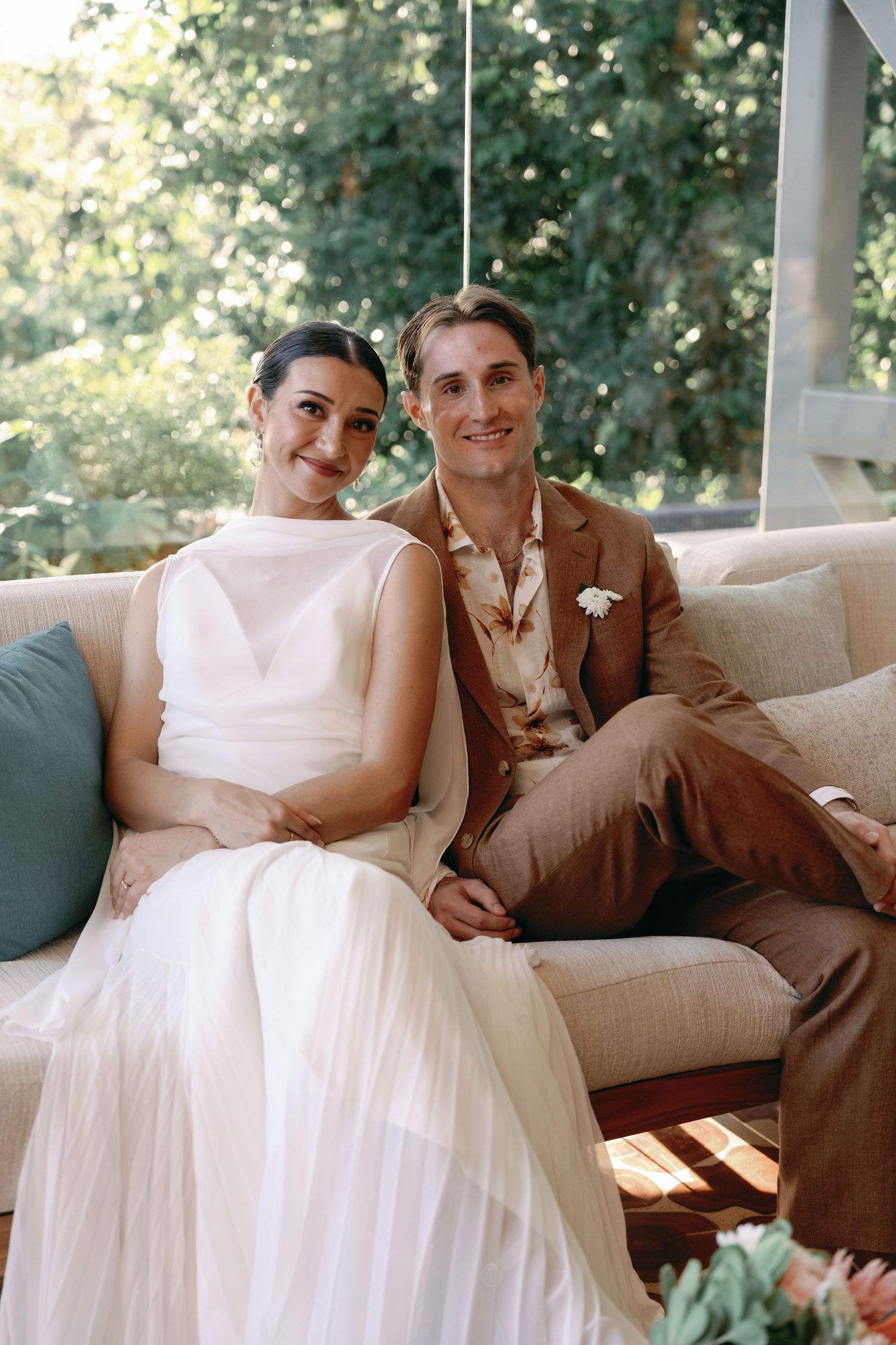 A couple relaxes together on a beige couch, with the bride in white and groom in brown suit in a sunlit setting.