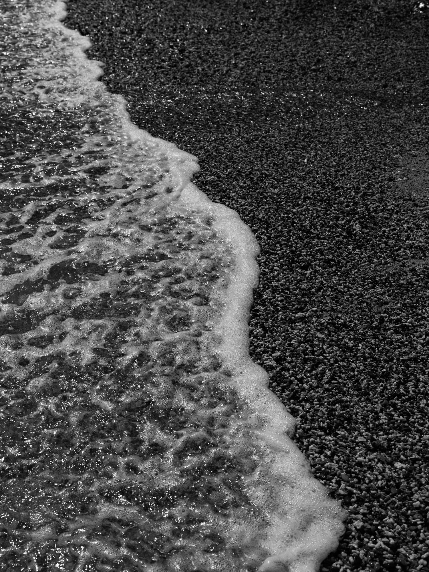 Black and white close-up of ocean waves breaking on a dark sandy beach.