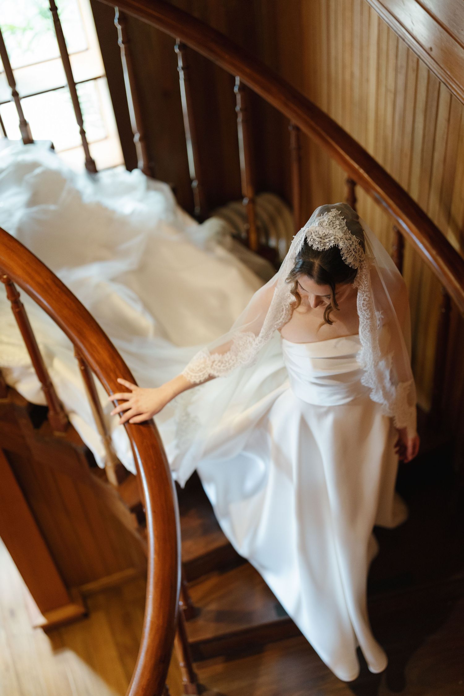 Bride in white gown and lace veil descends elegant wooden spiral staircase, her dress trailing behind her.