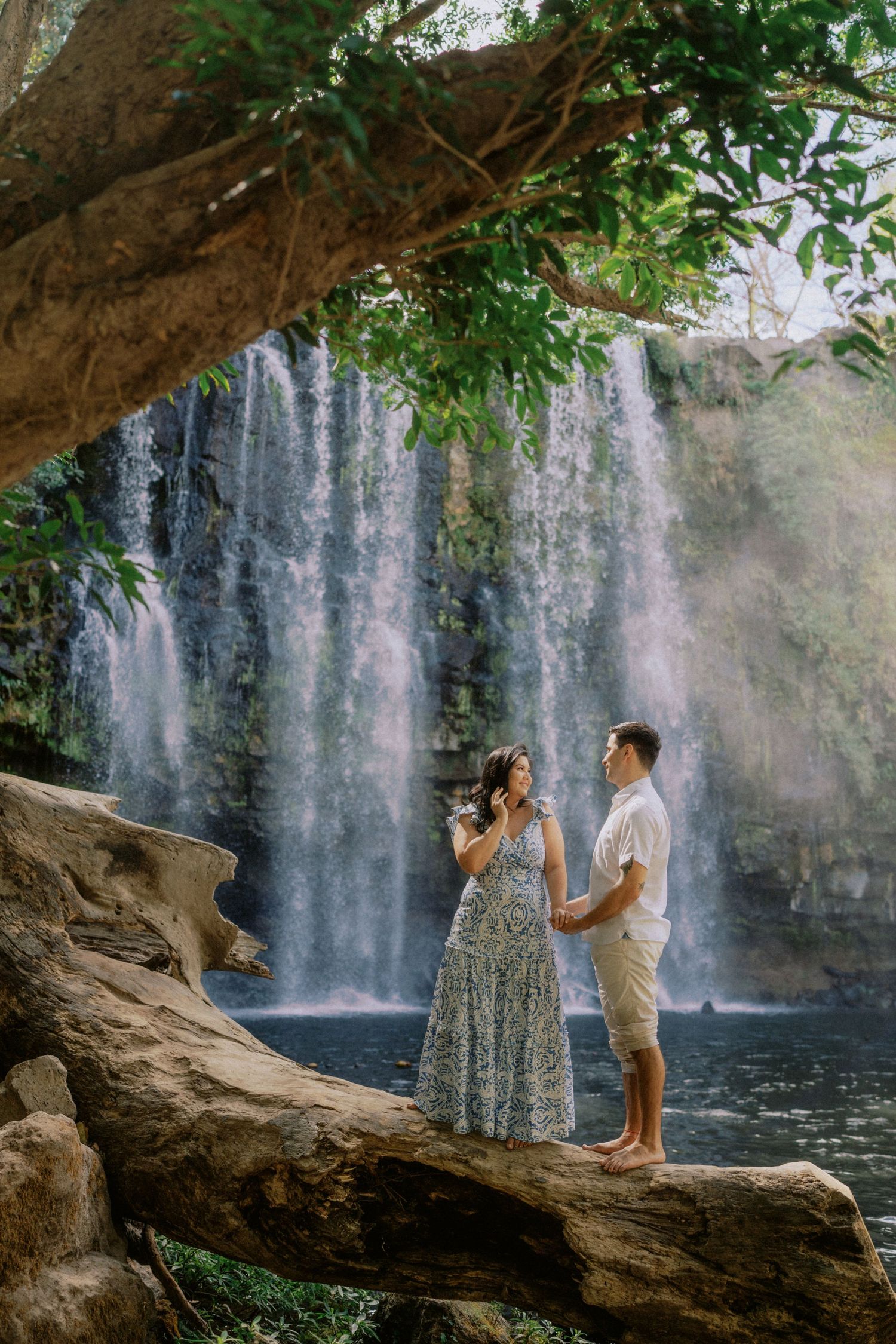 A couple shares a romantic moment on a fallen tree trunk with a waterfall backdrop.