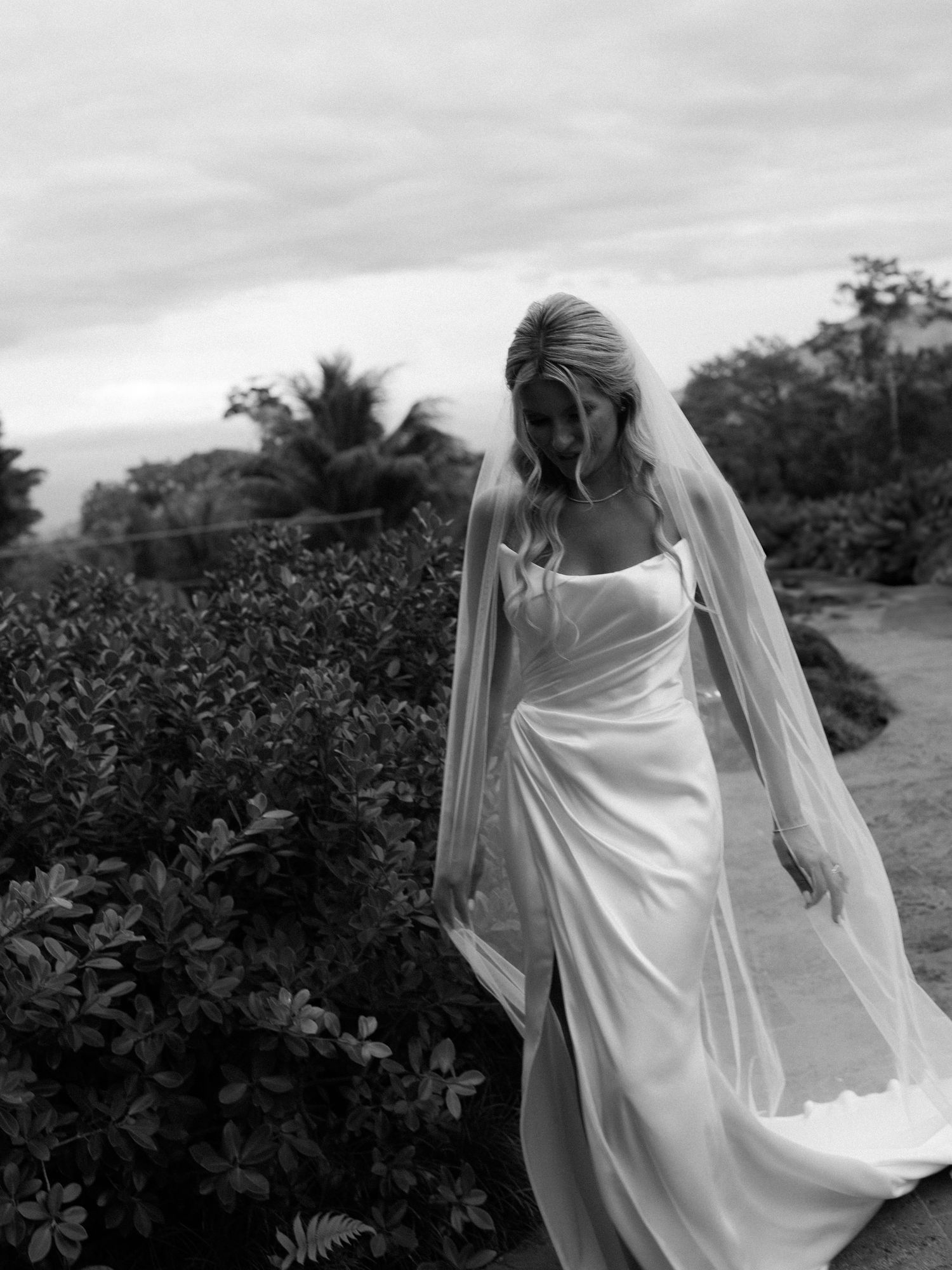 Bride in flowing white dress and veil walking through garden path.