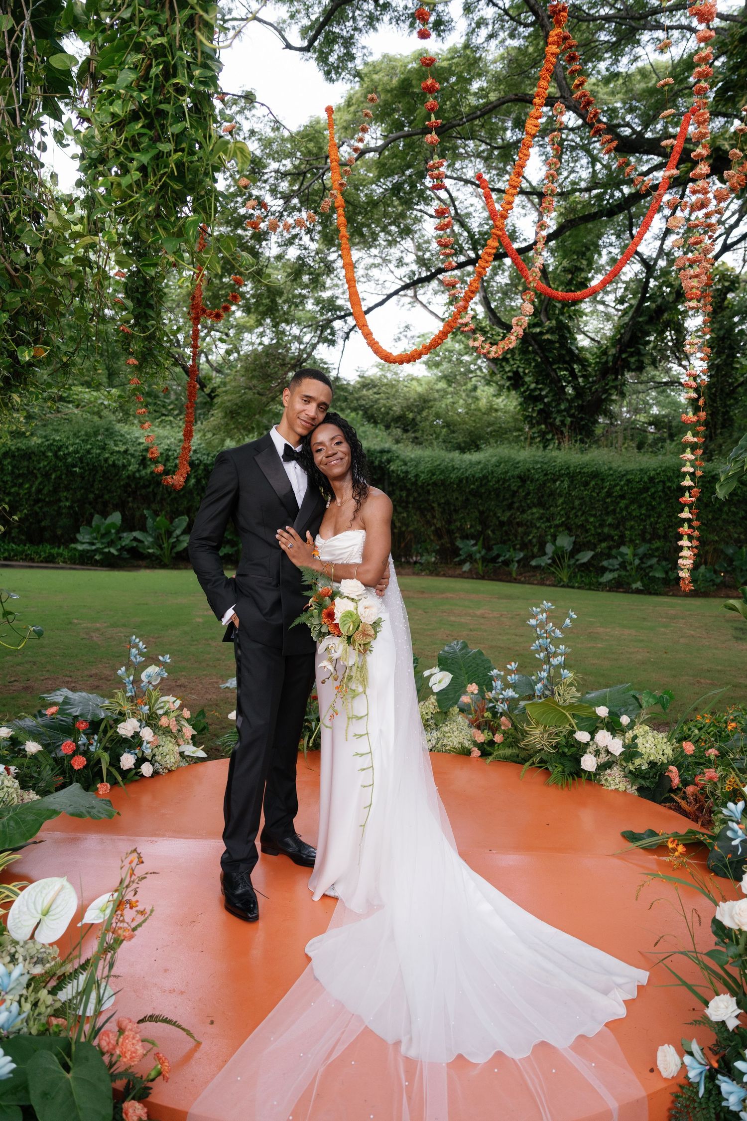 Wedding ceremony setup with coral-colored dance floor and hanging floral decorations in an outdoor garden setting.