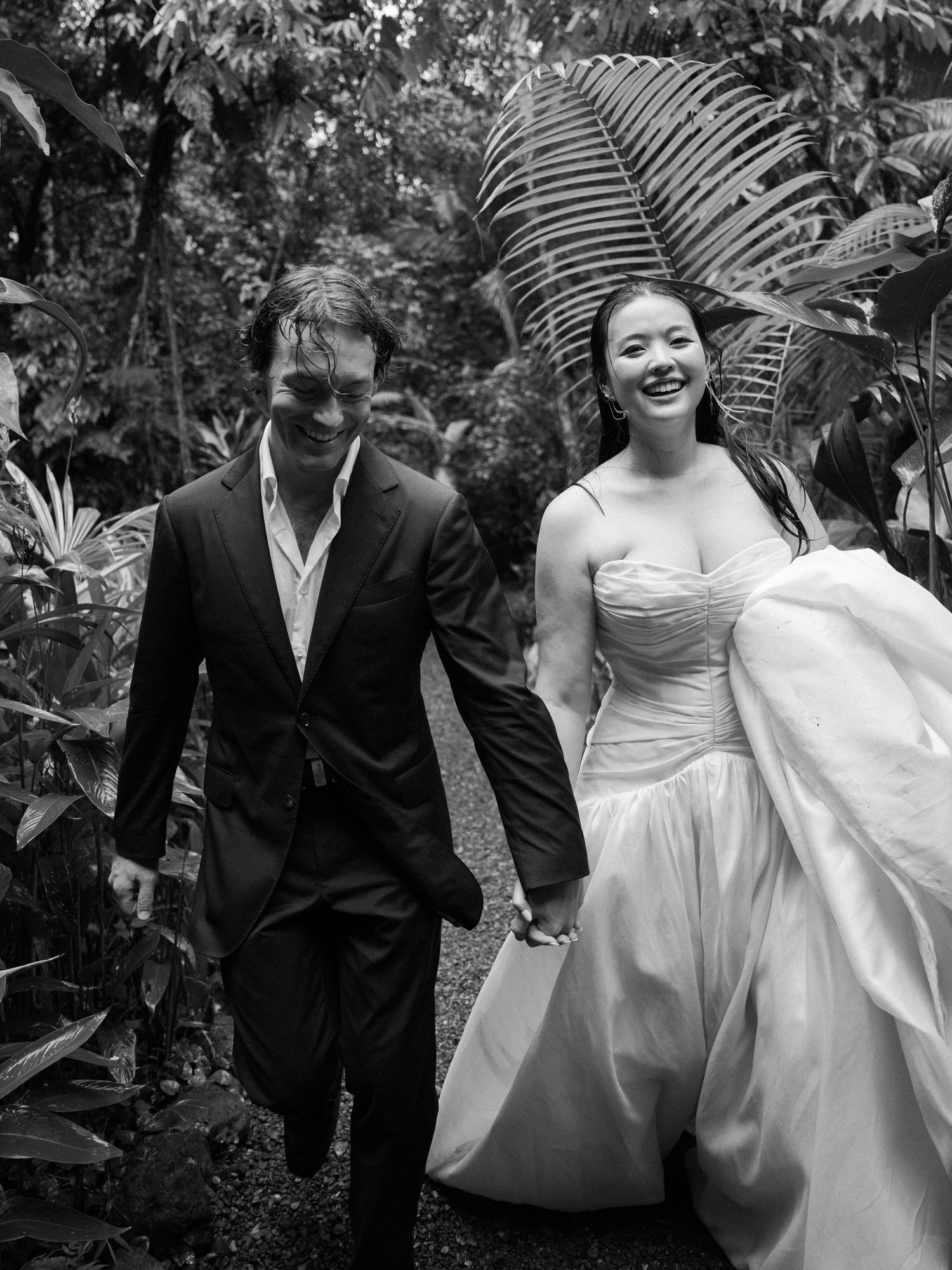 A joyful wedding couple walks hand in hand through a lush tropical garden setting.
