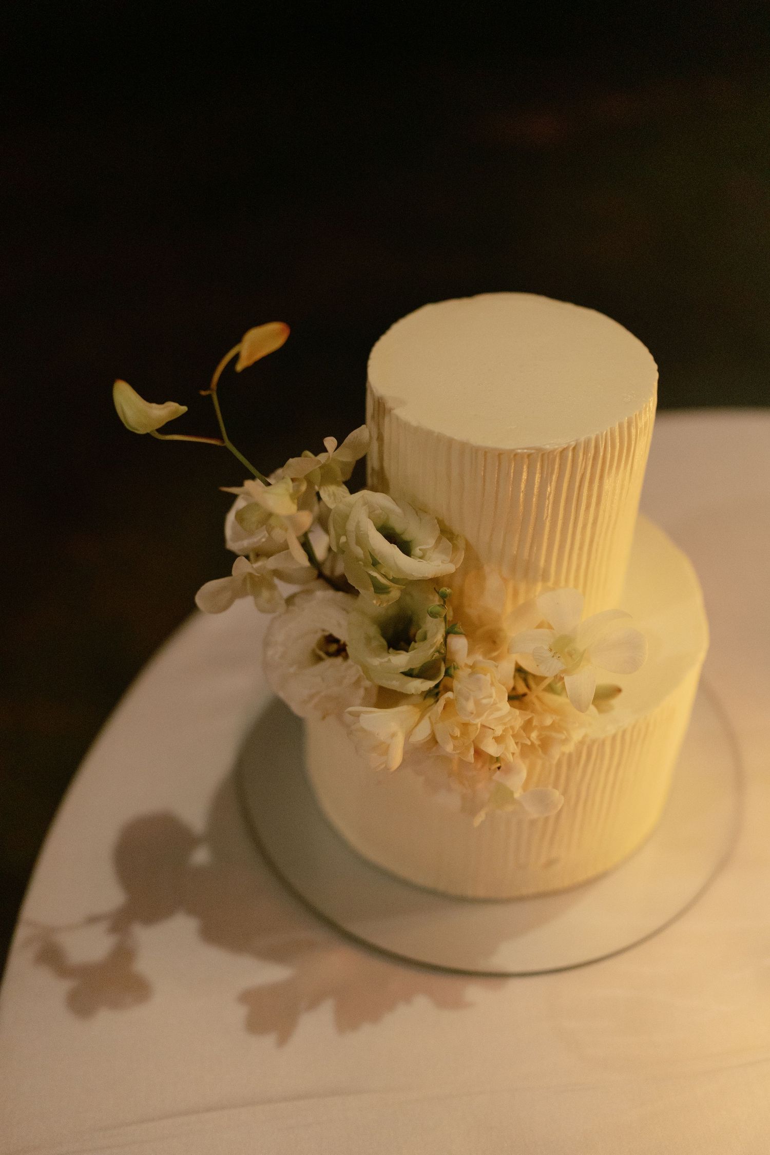 A simple white two-tiered wedding cake decorated with delicate white flowers and greenery on a light surface.