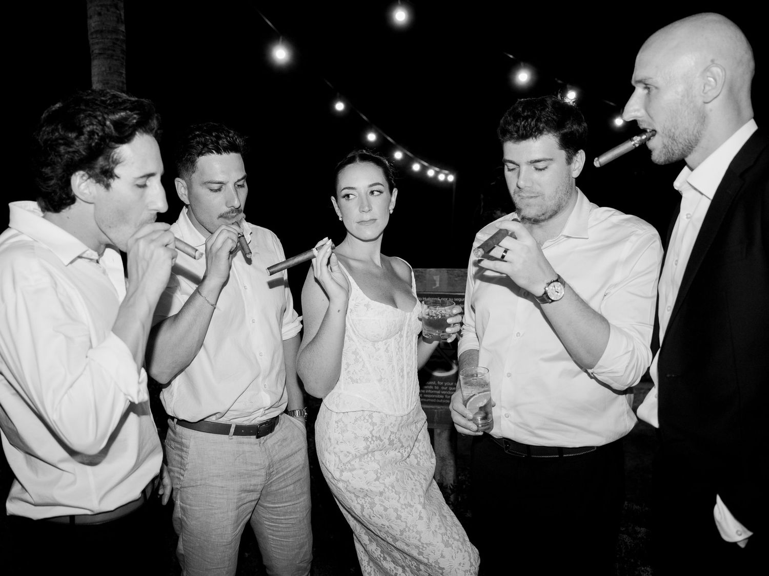 A vintage black and white photograph shows a group of people lighting cigarettes together at a nighttime gathering.