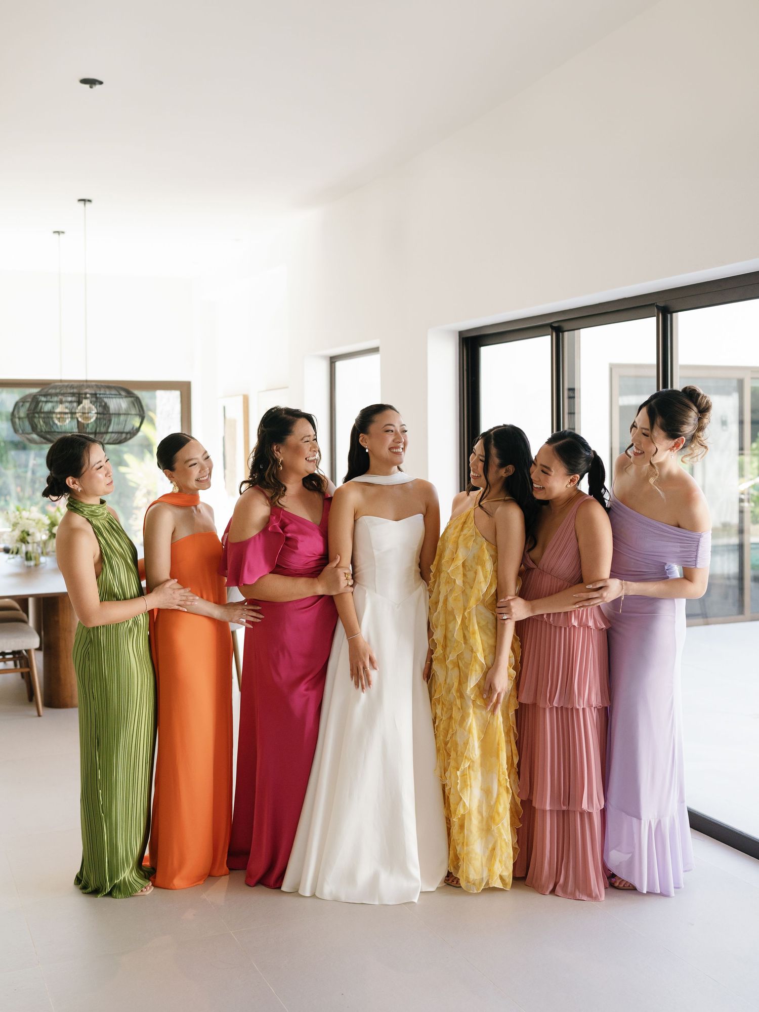 Bridesmaids wearing vibrant dresses in orange, green, pink, gold and purple pose together in a bright indoor space.