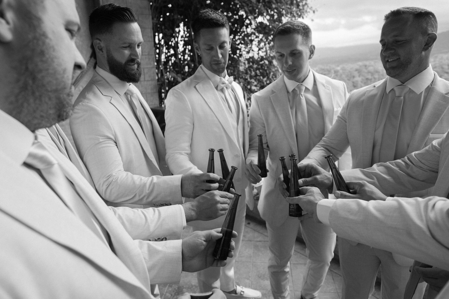 Groomsmen in white suits toast with drinks during a wedding celebration.
