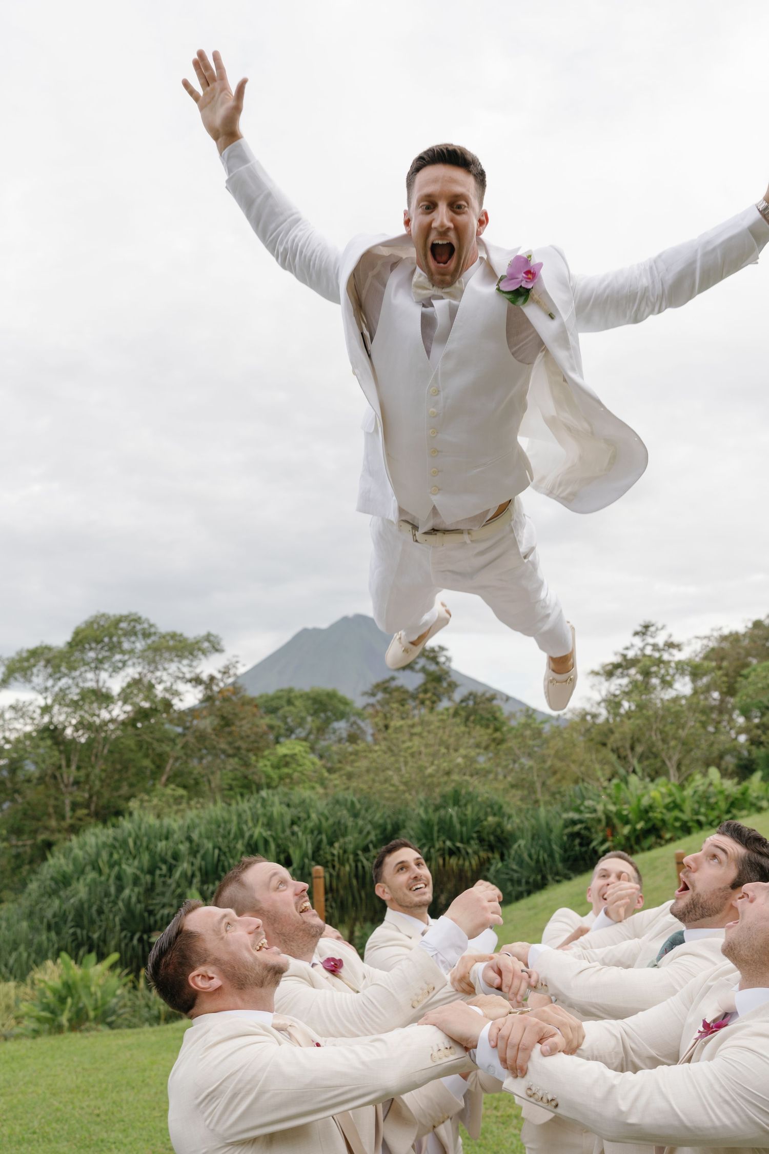 Groomsmen in white suits perform playful jumping photo on grassy hillside.