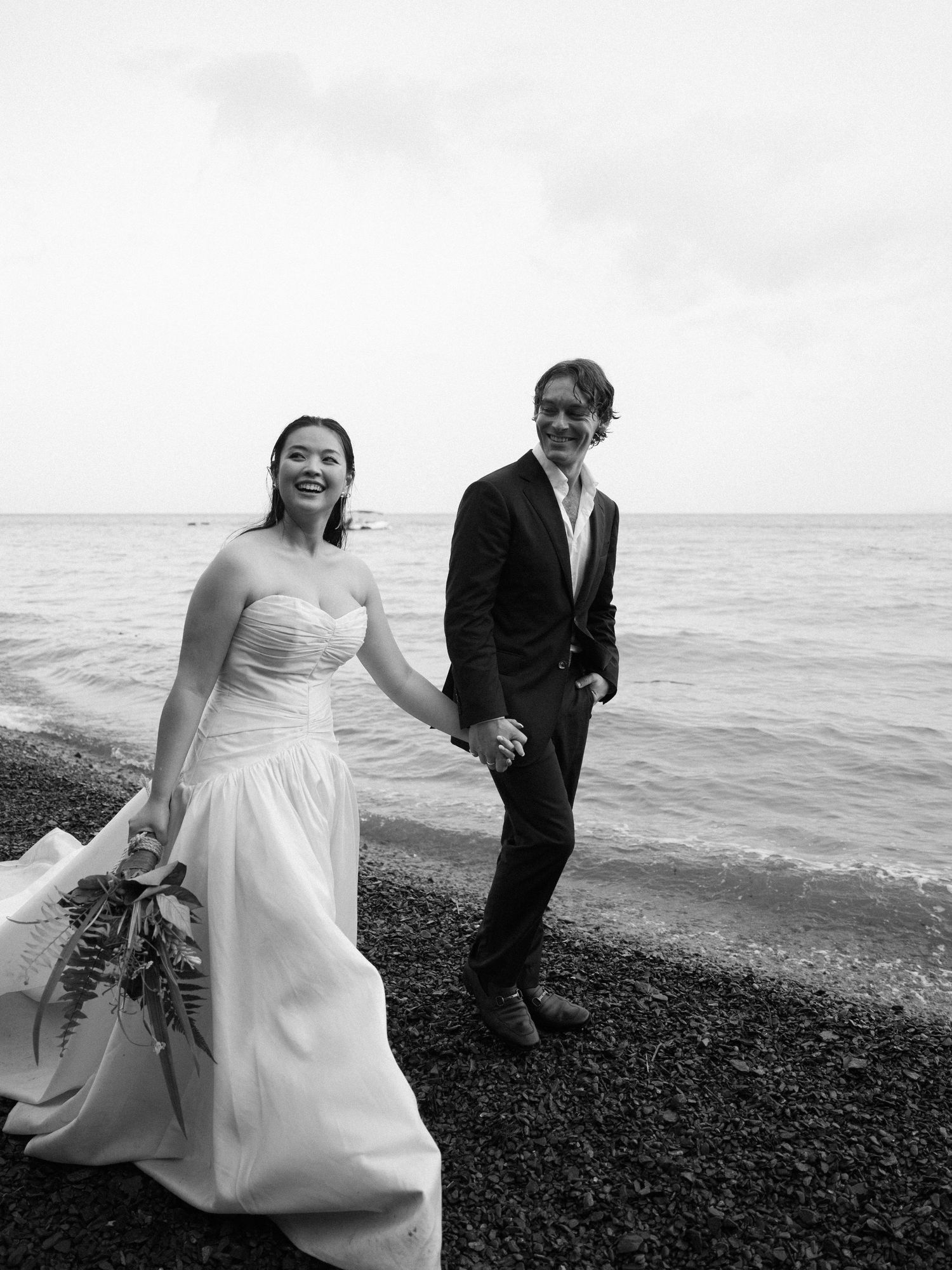 A couple in wedding attire poses on a rocky beach with ocean waves in the background.
