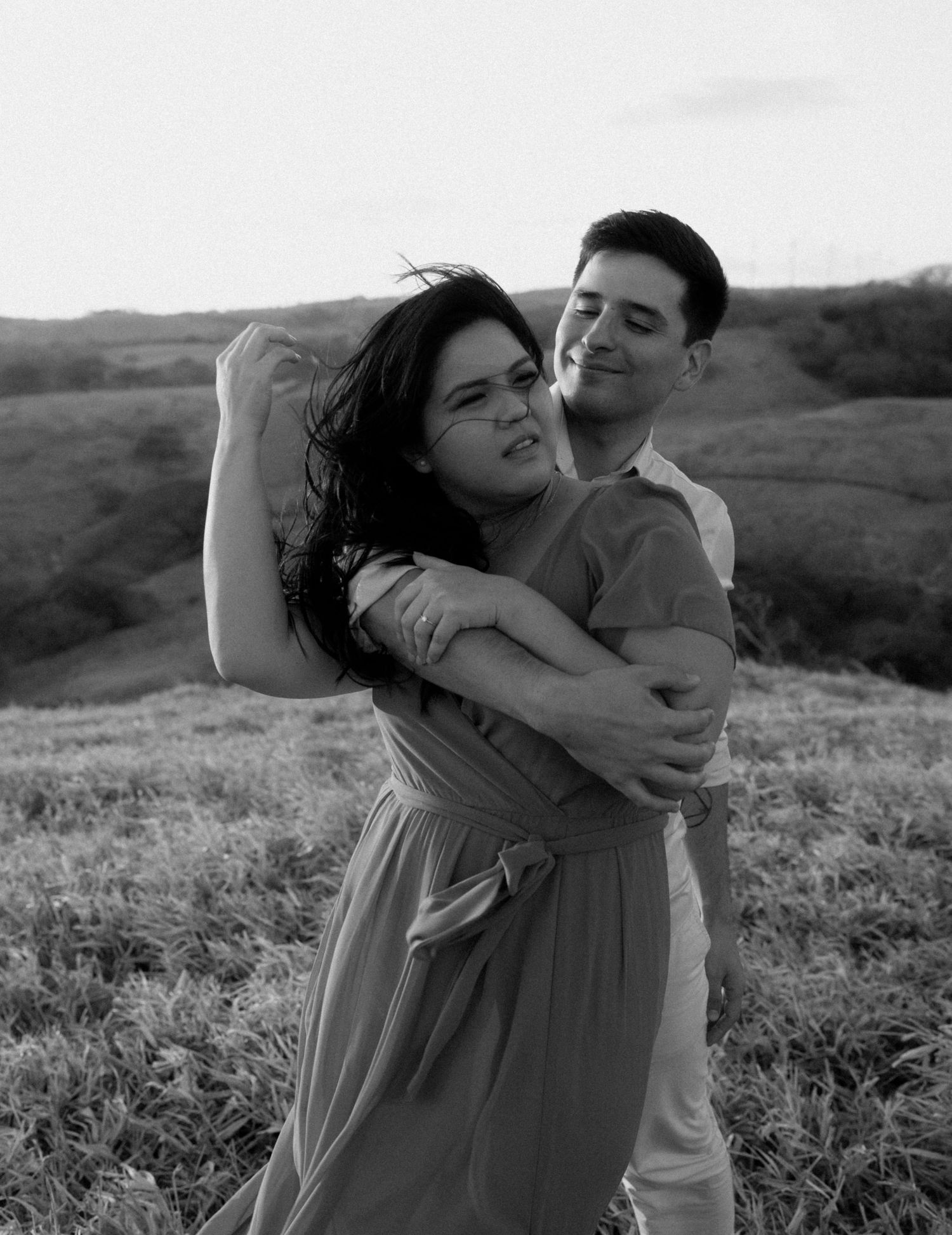 A romantic black and white portrait of a couple embracing in a hillside field.