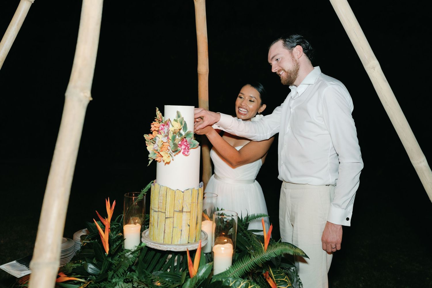 A couple celebrates their wedding with a tiered cake decorated with greenery and flowers at an outdoor reception.