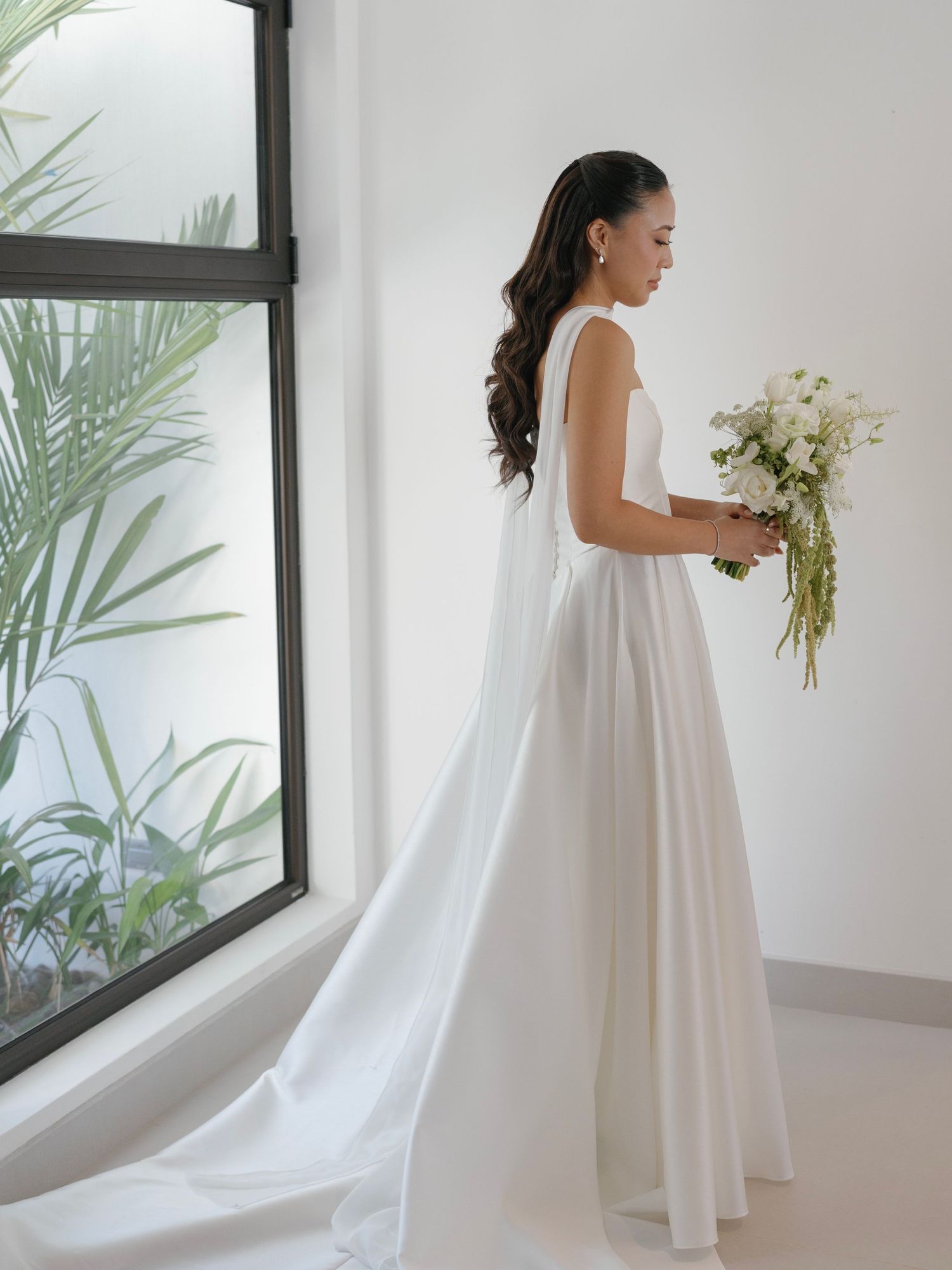 Elegant white wedding dress with flowing train in a bright modern room with palm leaves.