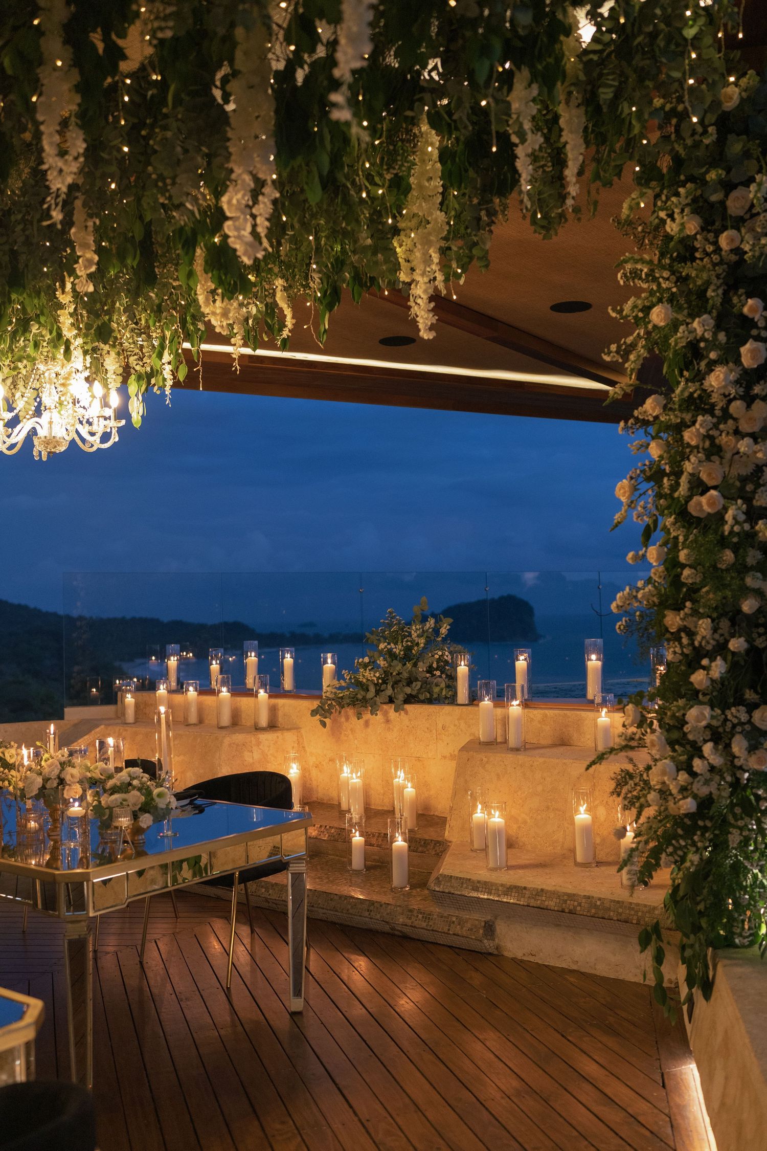 An elegant outdoor terrace decorated with hanging flowers and candles overlooking a twilight ocean view.