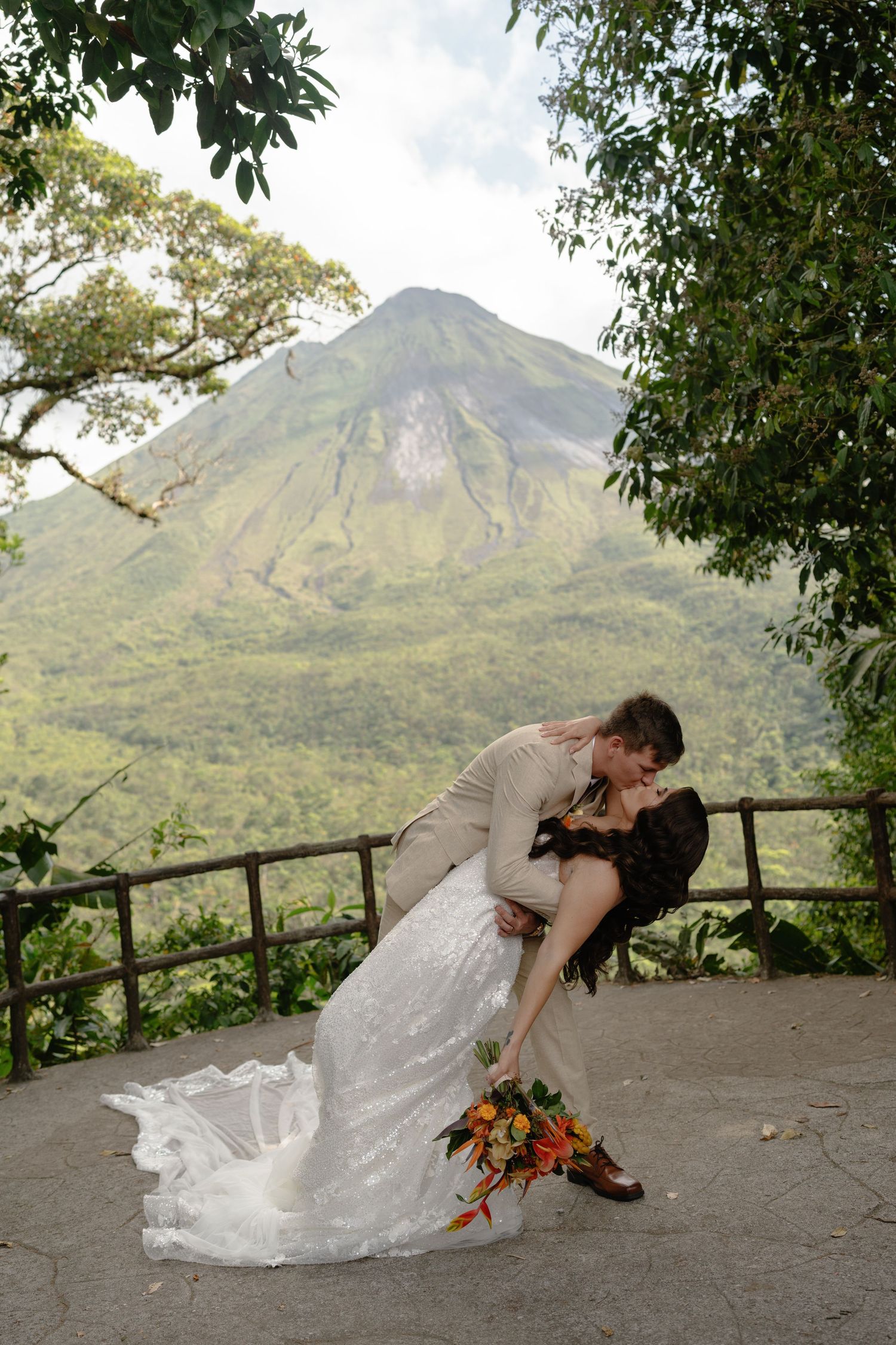 A couple in wedding attire dances playfully against a mountain backdrop with tropical foliage.