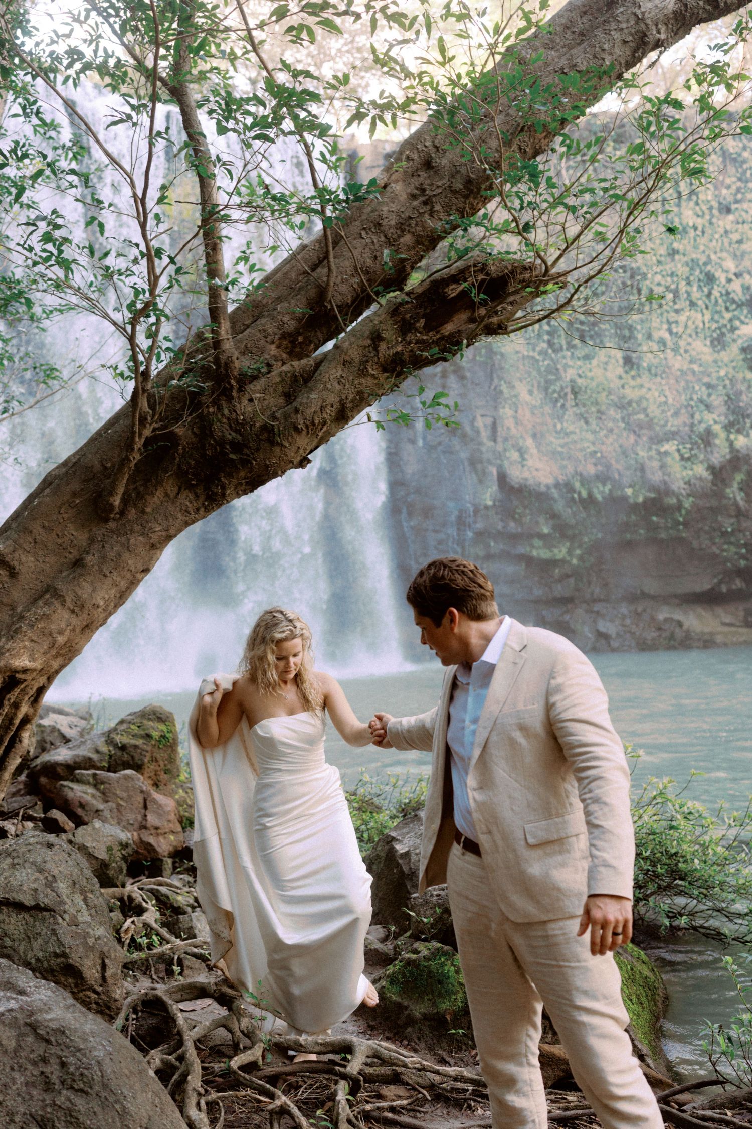 Two people in white outfits walk carefully along mossy rocks near a waterfall.